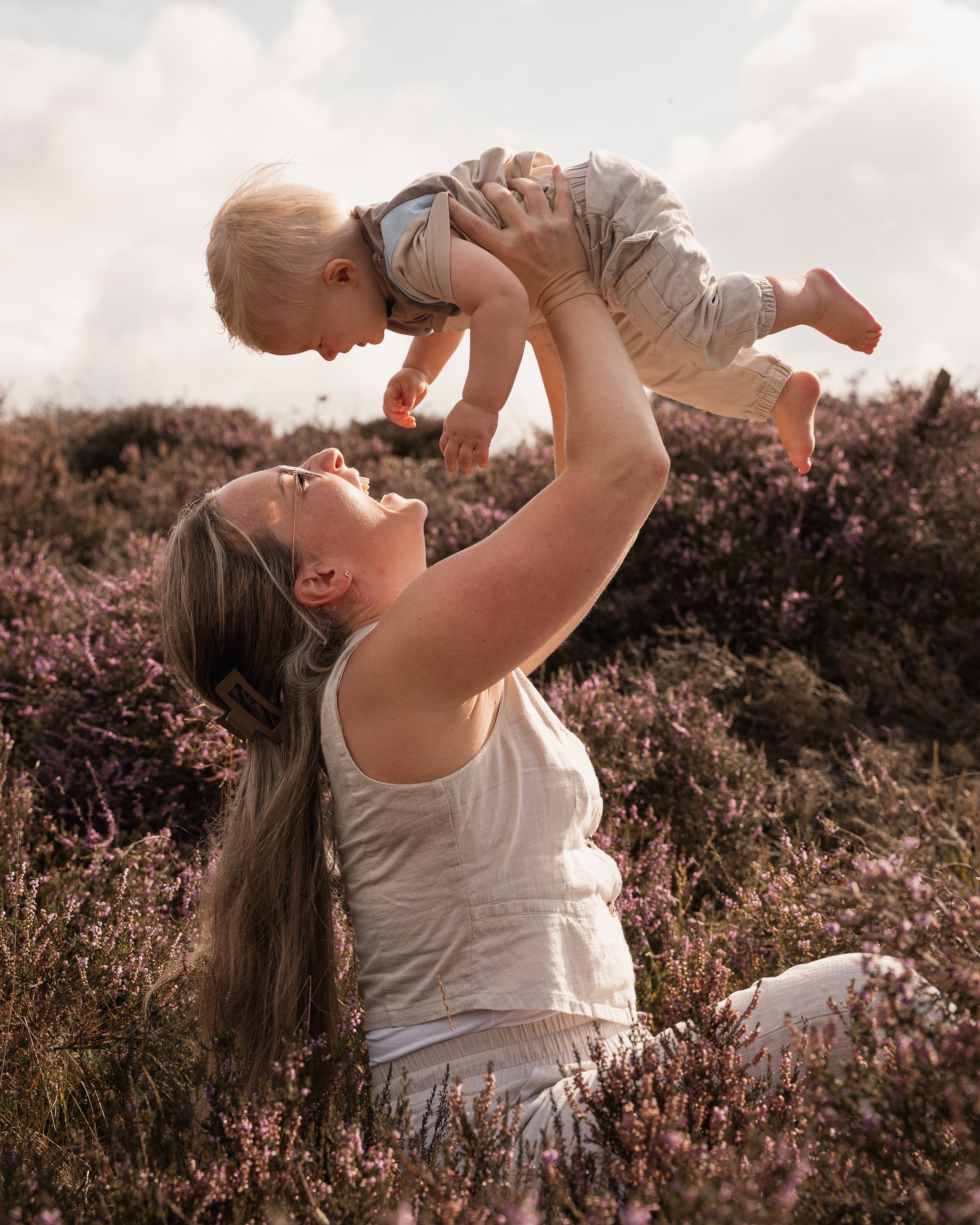 Een vrouw die in een veld met paarse bloemen zit, houdt een jonge jongen omhoog, terwijl hij in de lucht ligt.