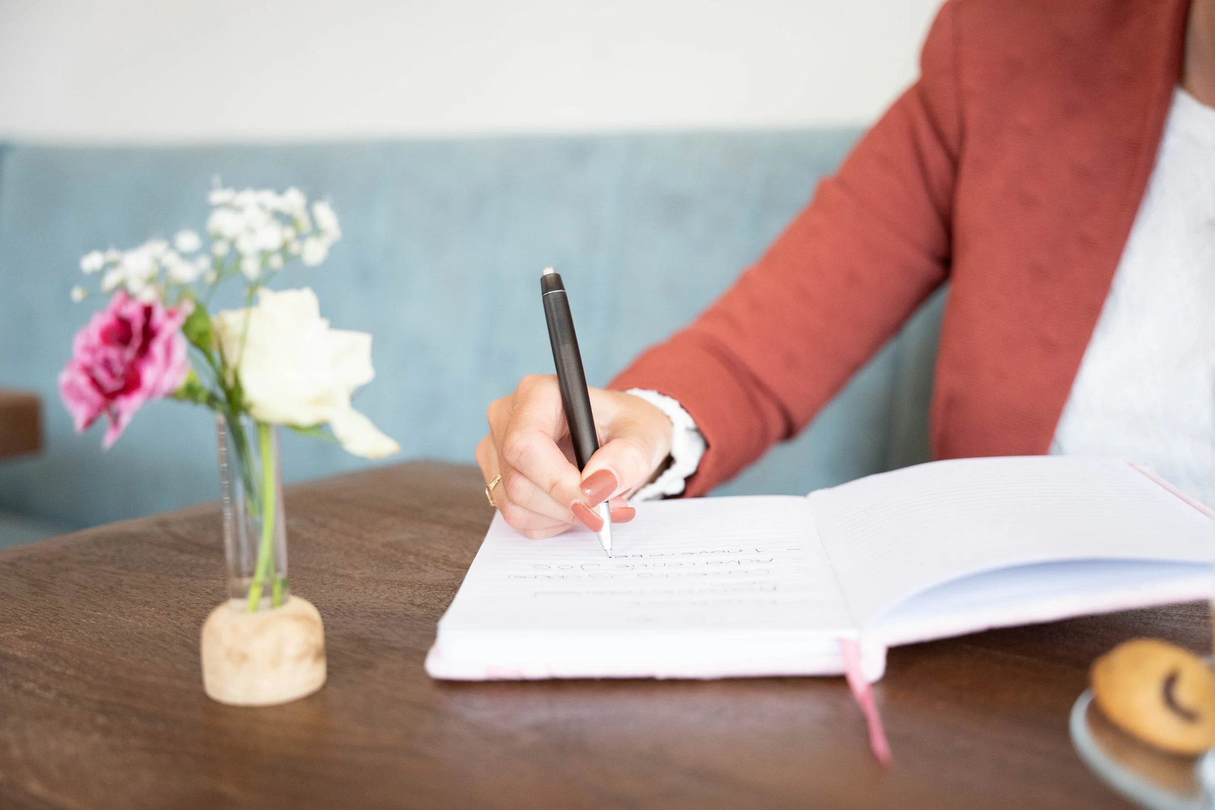 Persoon schrijft in een open notitieboekje op een houten tafel, naast een bloemstuk in een korte vaas met witte en roze bloemen en een schuimrubber onderkant, en een koekje op een bord.