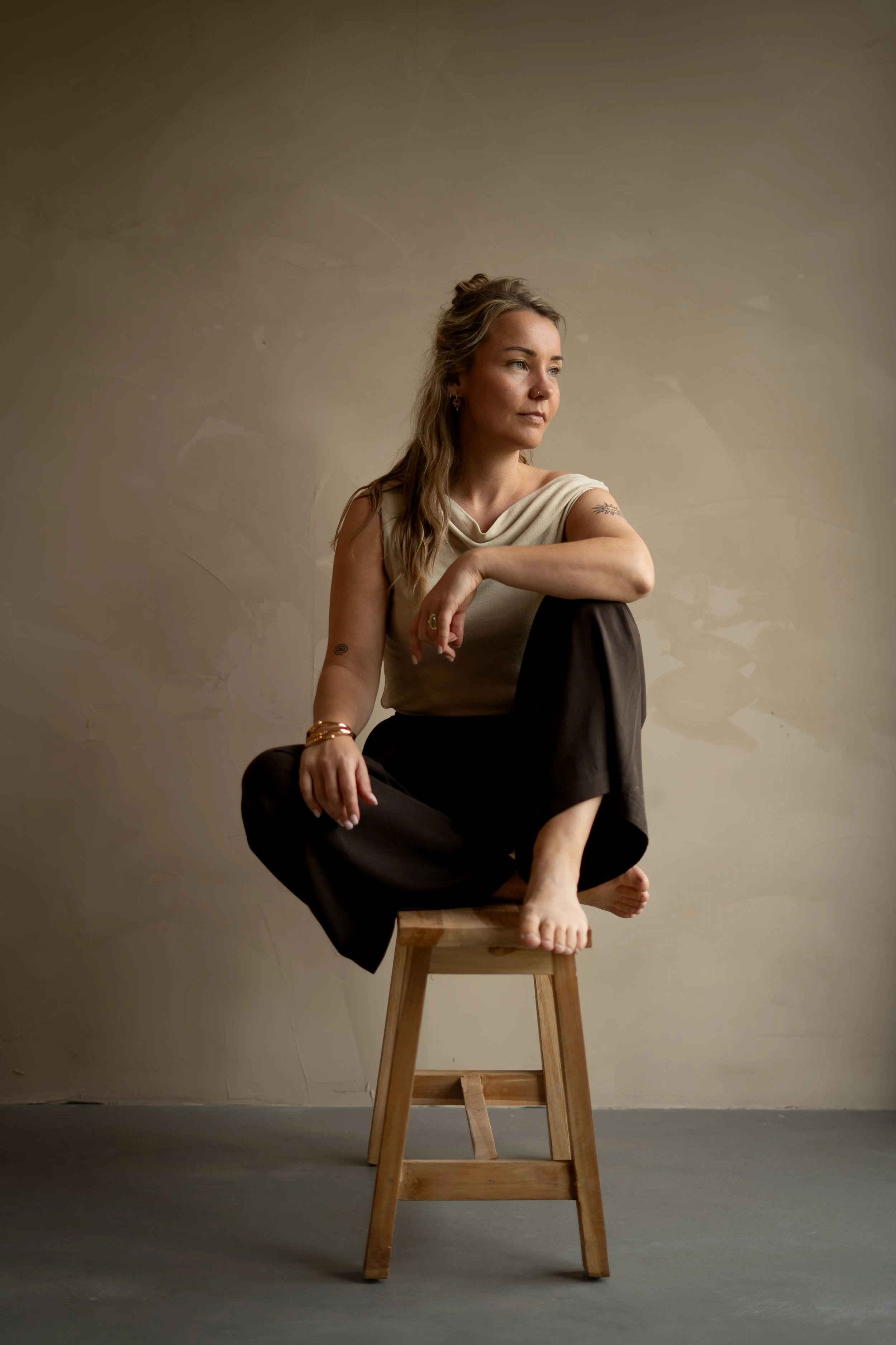 Vrouw met curly haar en bril, gekleed in een wit overhemd en lichte spijkerbroek, die een laptop vasthoudt, staat in een lichte kamer met planten en een houten plank.