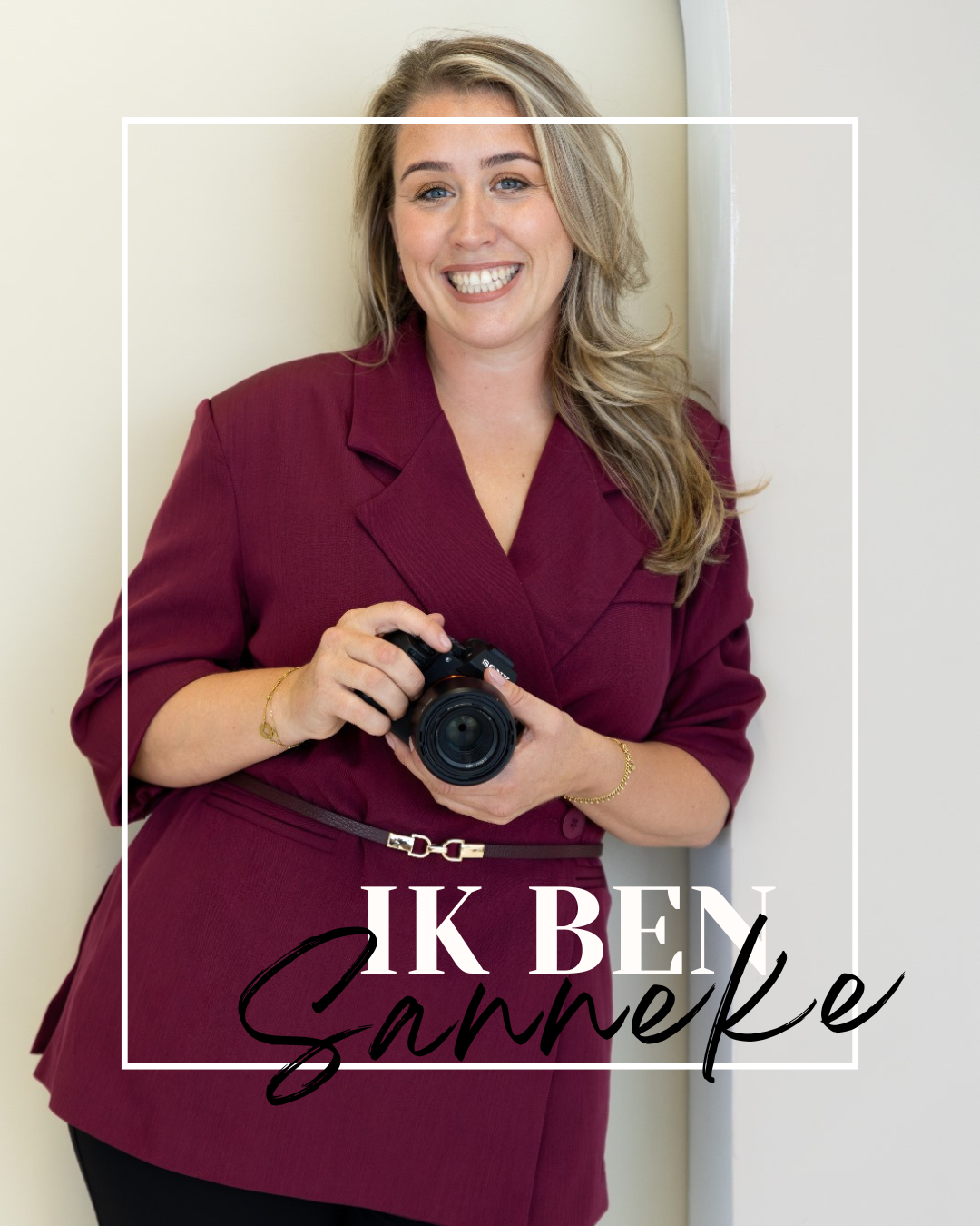 Vrouw met lang, blond haar, glimlachend, in een rokkenpak die een camera vasthoudt, leunend tegen een muur.