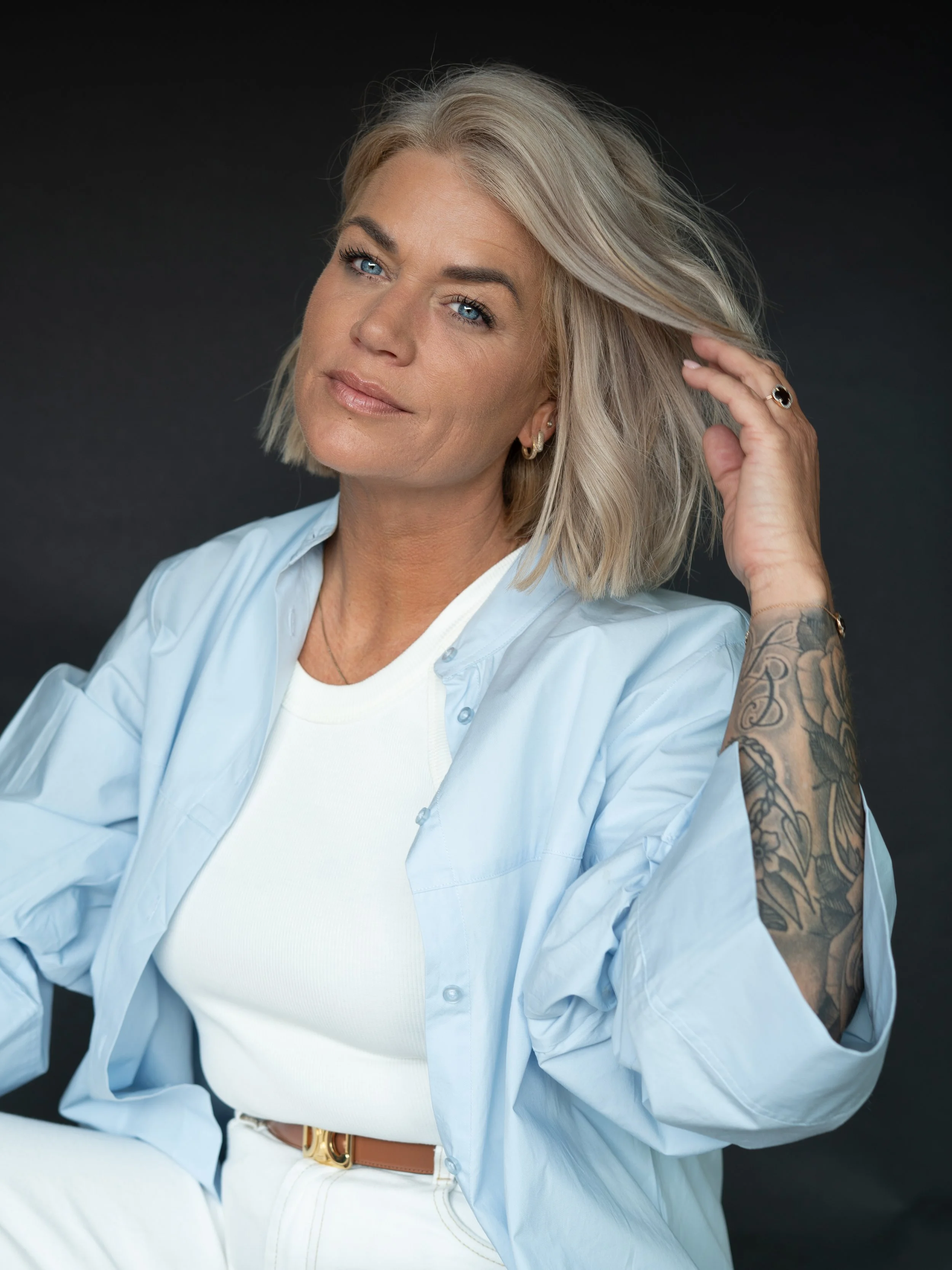 Portret van een vrouw met blonde, schouderlange haar die de ene hand door haar haar haalt, gekleed in een blauwe blouse en witte top, tegen een donkere achtergrond.