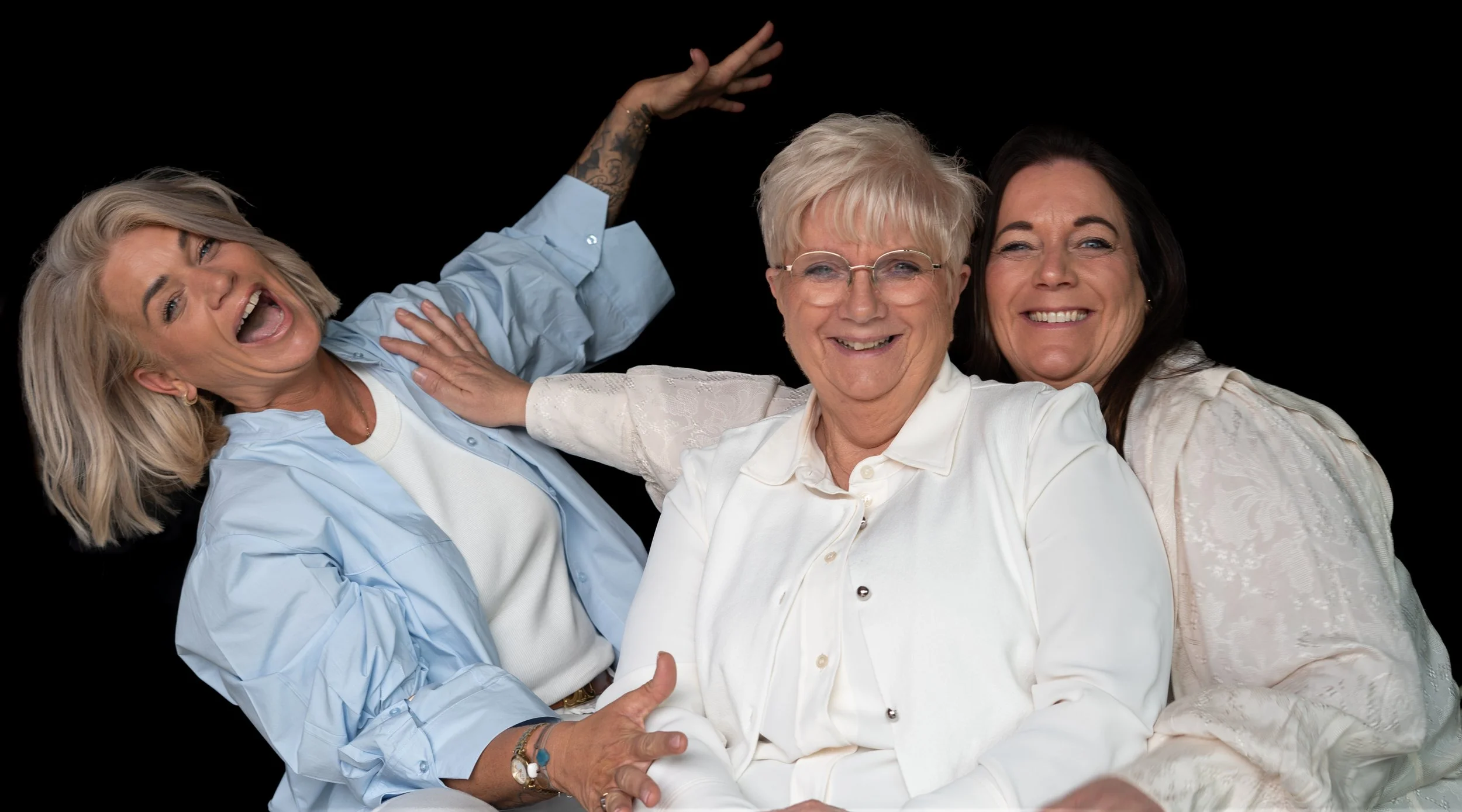 Vier lachende vrouwen, waarvan één oudere vrouw in het midden en drie jongere vrouwen eromheen, poseren tegen een zwarte achtergrond.