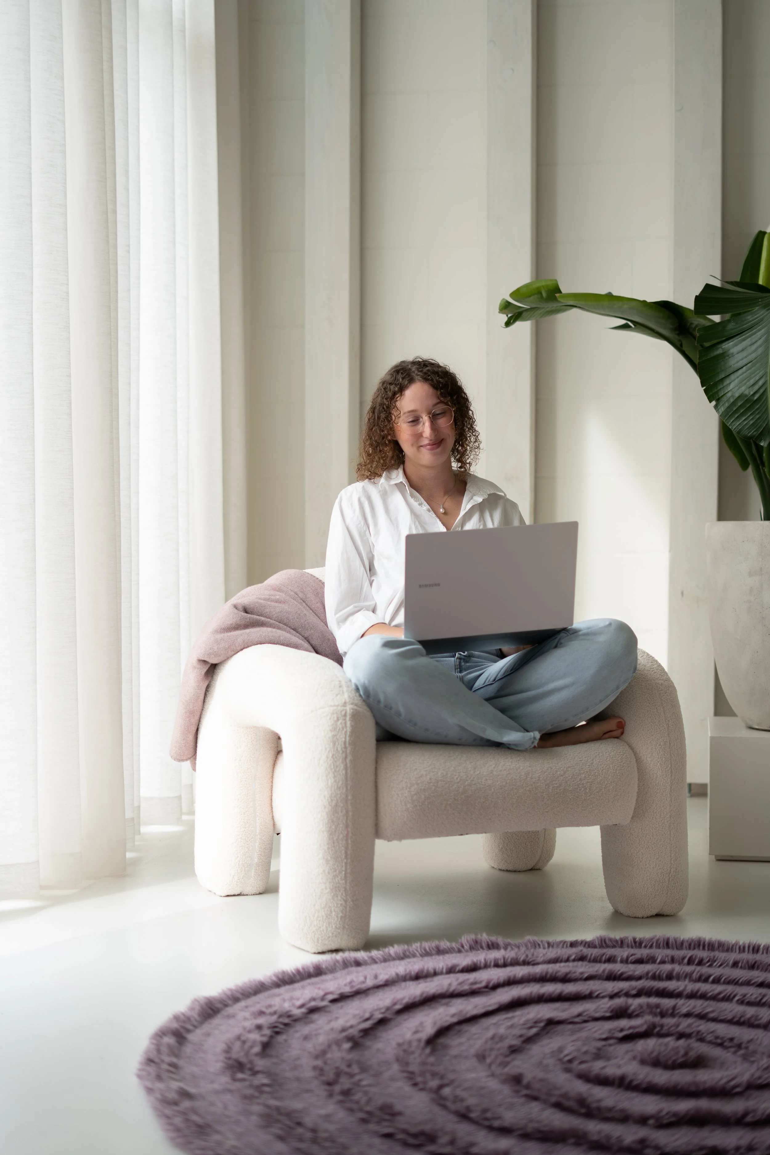 Vrouw zit op een beige gedraaide stoel met haar benen gekruist en gebruikt een laptop, in een lichte kamer met grote witte gordijnen en een groen plant.