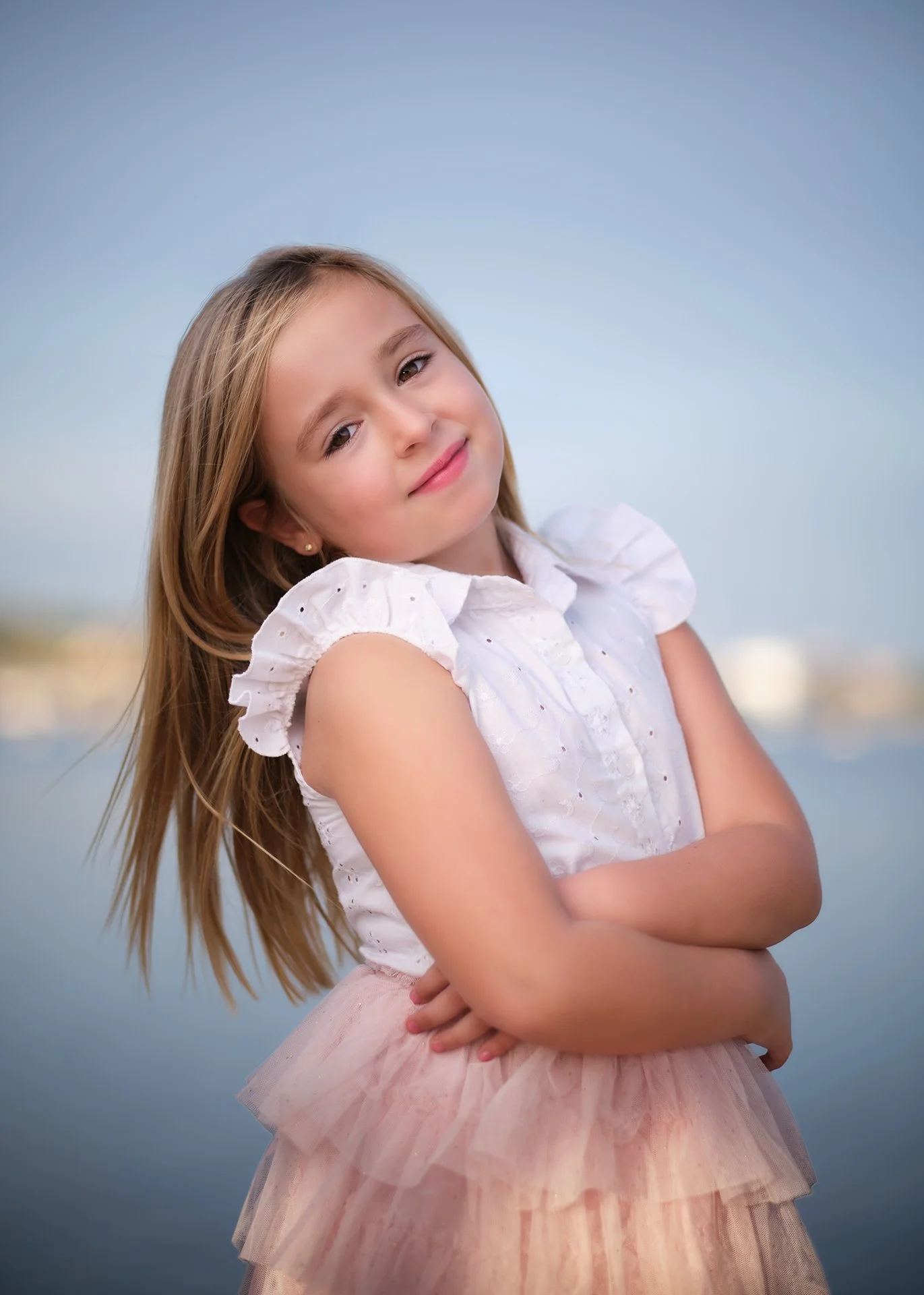Retrato de una niña con cabello largo rubio, levantada con un vestido blanco con volantes en los hombros y una falda de tul rosa, posando frente a un fondo con agua y cielo despejado.