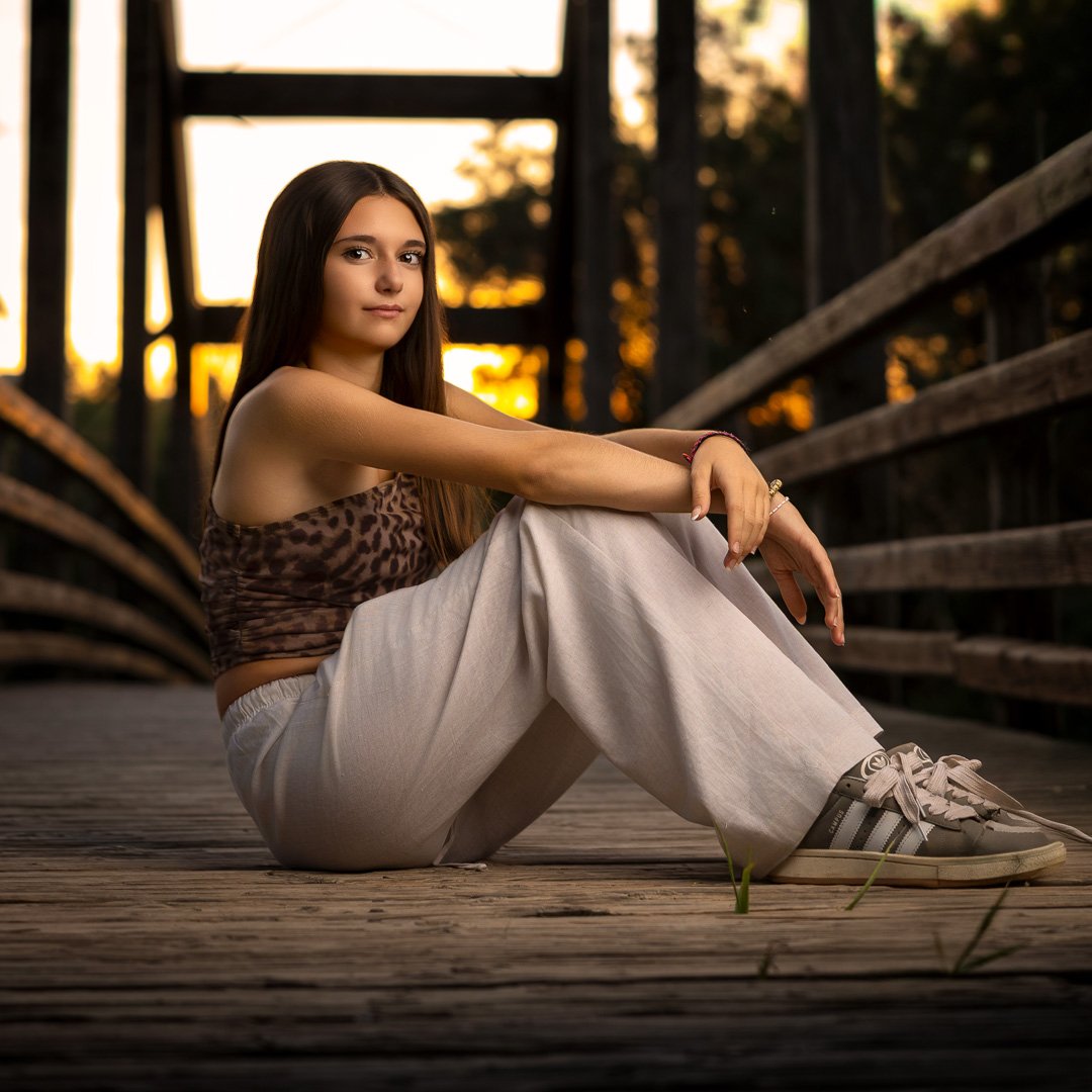 Joven mujer sentada en un puente de madera al atardecer, con vestimenta casual y cabello largo, mirando hacia la cámara.