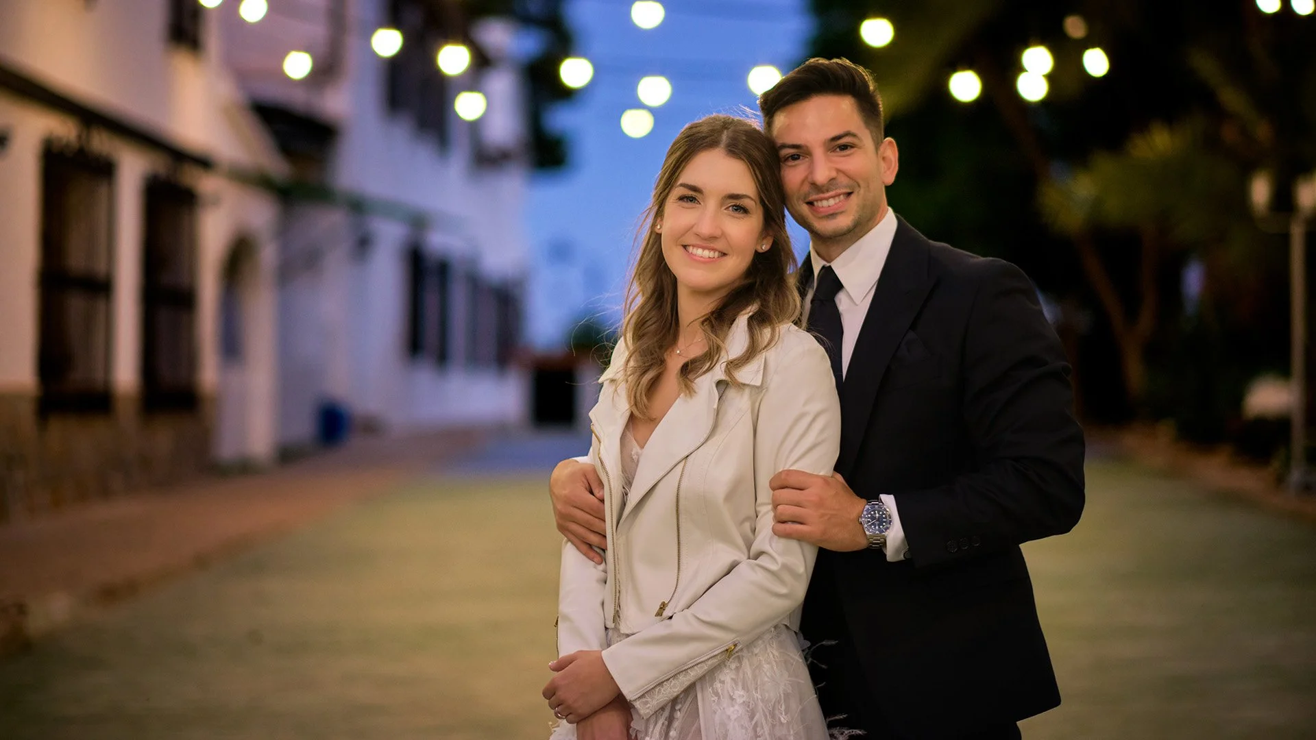 una pareja sonriendo en una calle iluminada por luces durante la noche, con árboles y edificios de fondo.