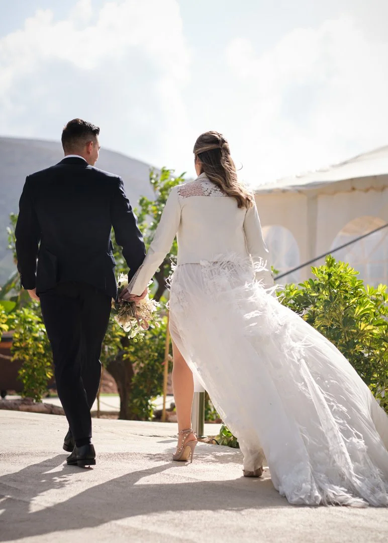 Pareja vestida de boda, el hombre con saco oscuro y la mujer con vestido largo y blusa blanca, caminando de la mano en un espacio al aire libre.