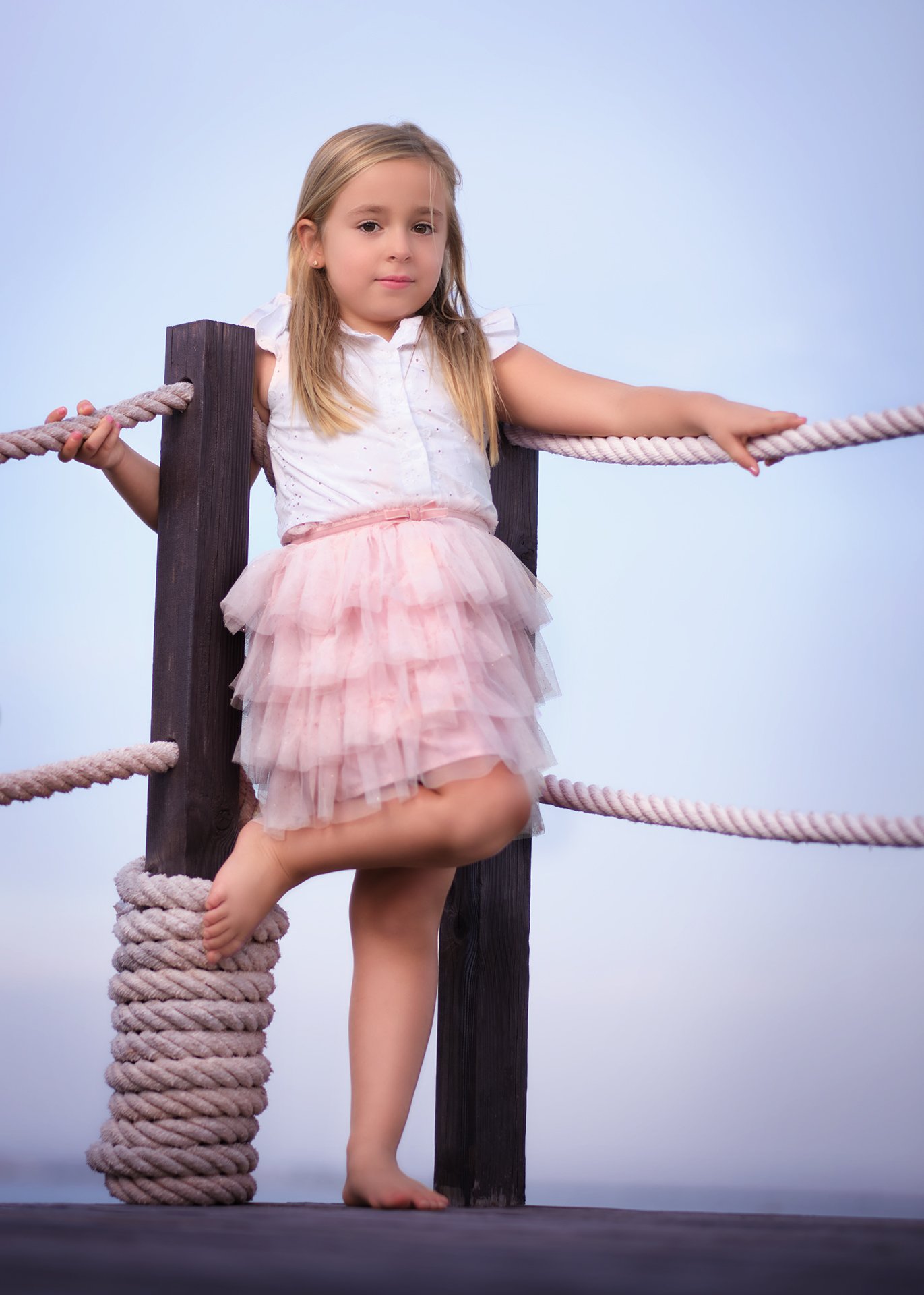 Niña con vestido rosa y blusa blanca, apoyada en una cuerda de un puente de madera, con una pierna levantada y el pie apoyado en la cuerda, fondo azul claro.