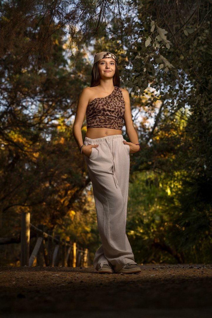 Joven mujer con boina, top de leopardo, pantalones beige y zapatos deportivos, posando en un camino rodeado de árboles con follaje otoñal.