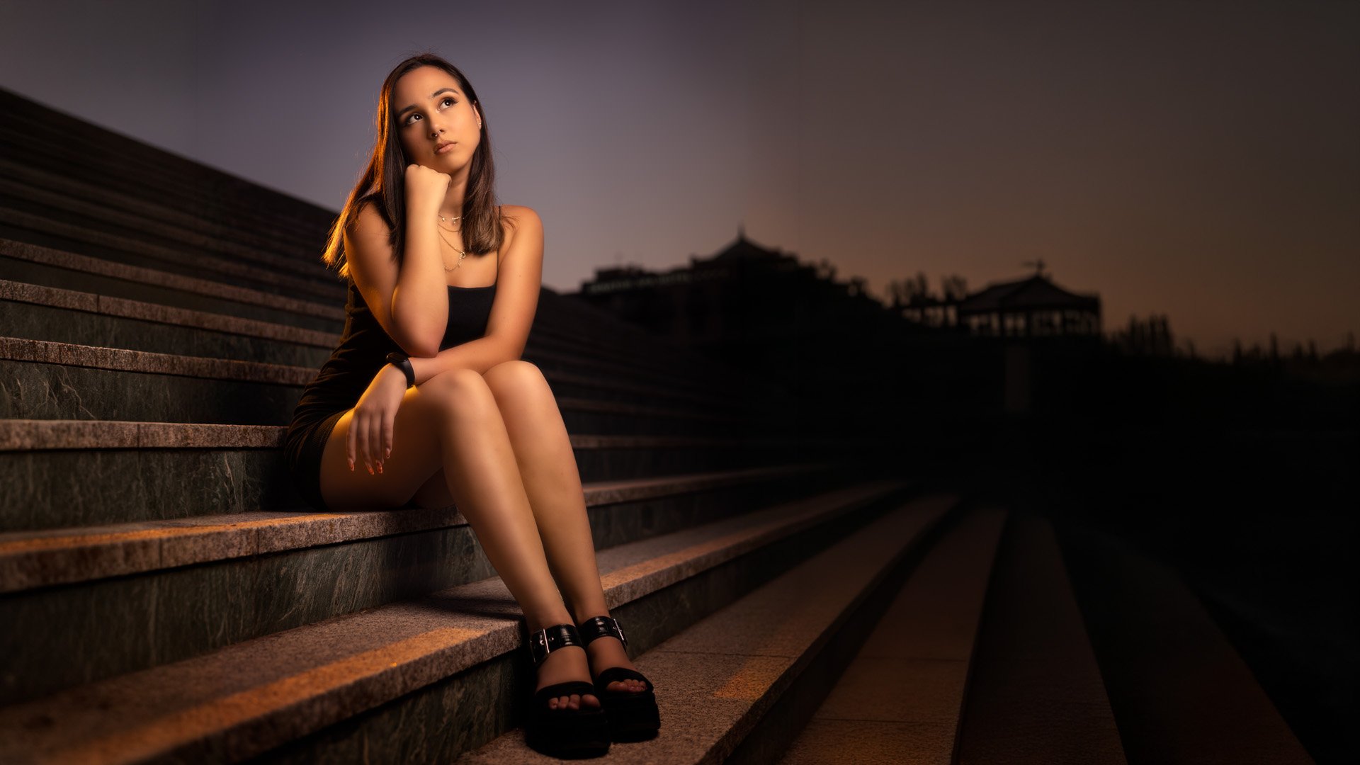 Joven mujer sentada en escaleras de noche, con expresión pensativa, luciendo vestido negro y calzado de glamour.