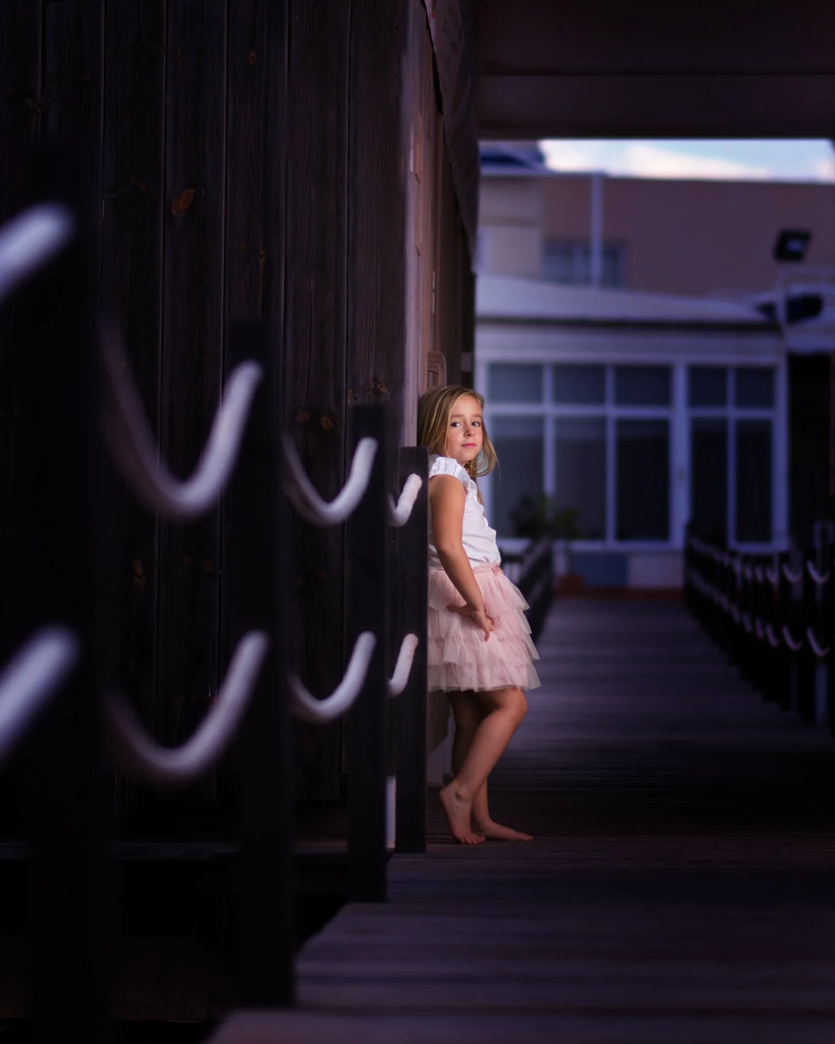 Una niña con vestido rosa y camiseta blanca se recarga en una pared de madera, en un pasillo exterior de un edificio, durante el atardecer.