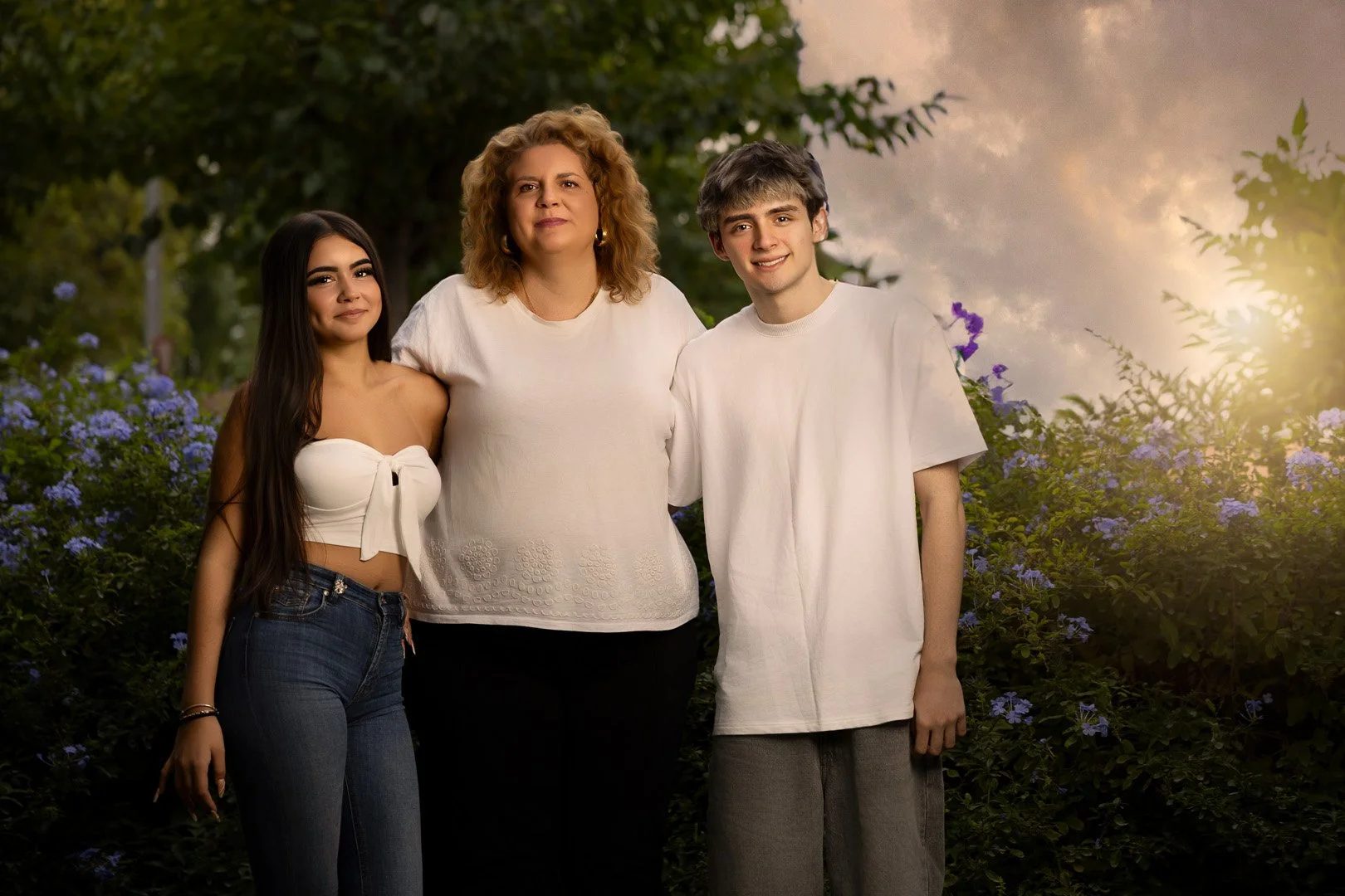 Una familia de tres personas, madre y dos adolescentes, posando al aire libre en un entorno con flores y árboles durante el atardecer.