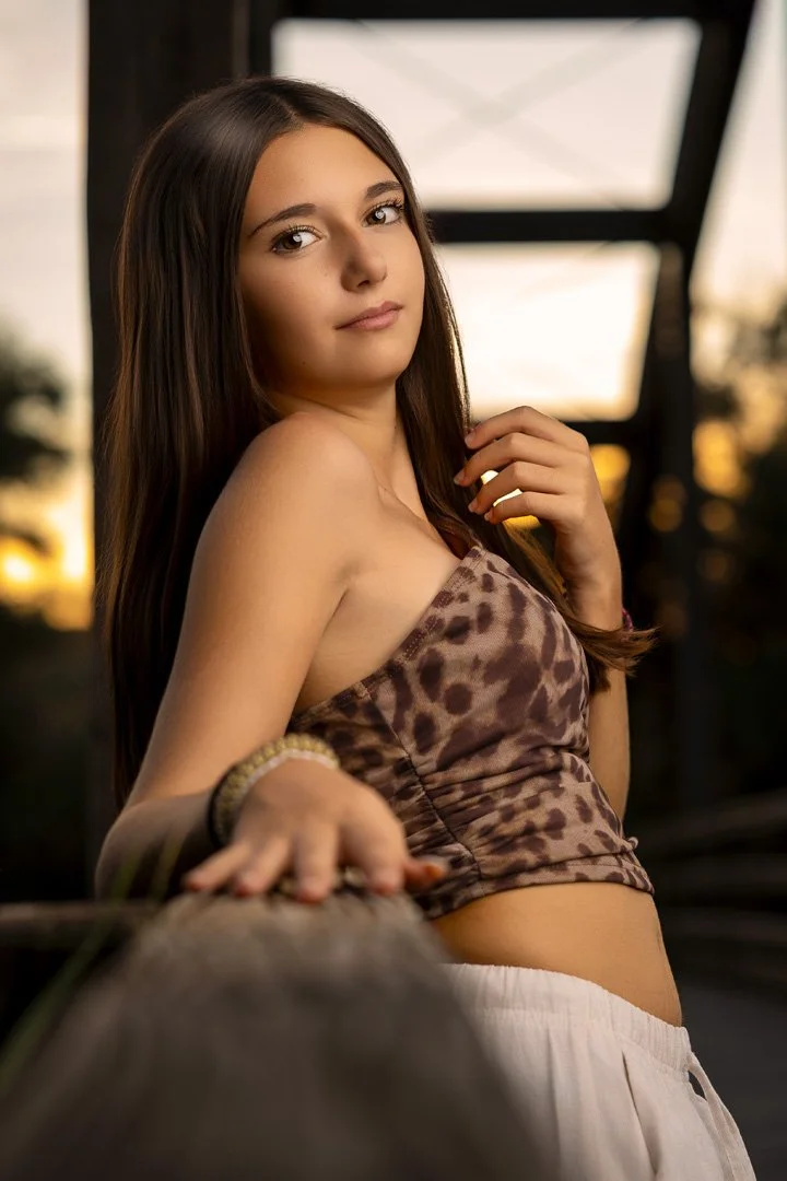 Joven mujer con cabello largo y liso, usando un top de animal print y pantalones claros, posando al atardecer con fondo difuso y estructura metálica.