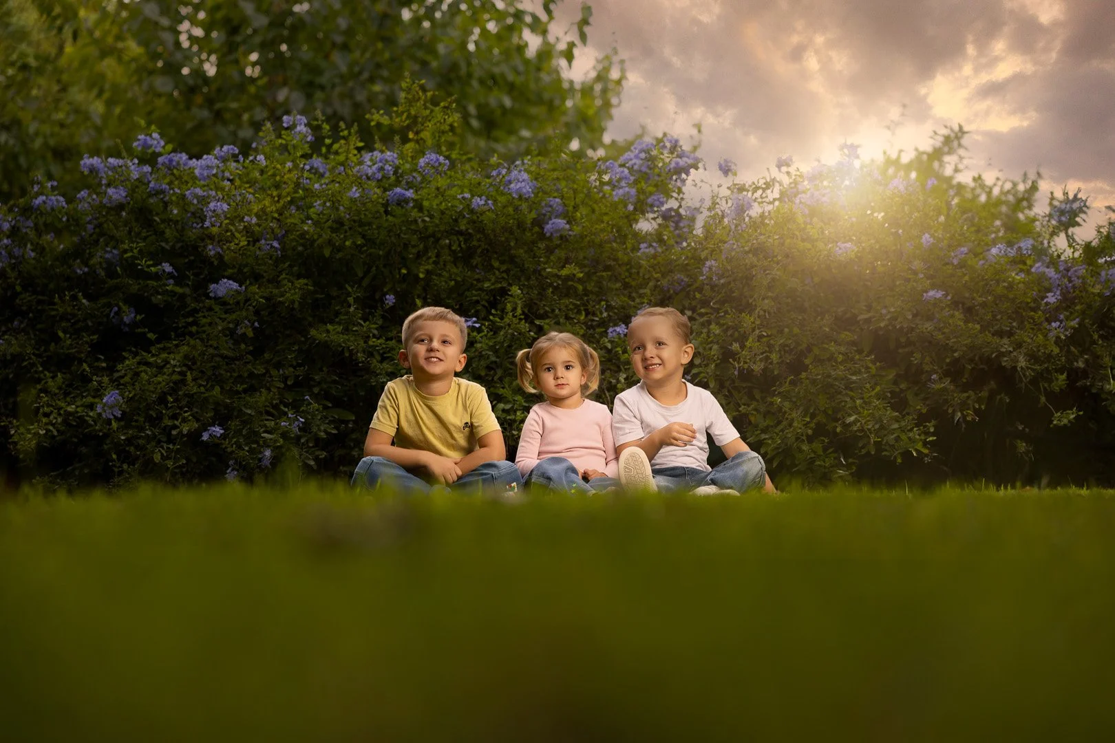 Tres niños pequeños sentados en el césped frente a arbustos con flores moradas, al atardecer con cielo nublado y sol brumoso.