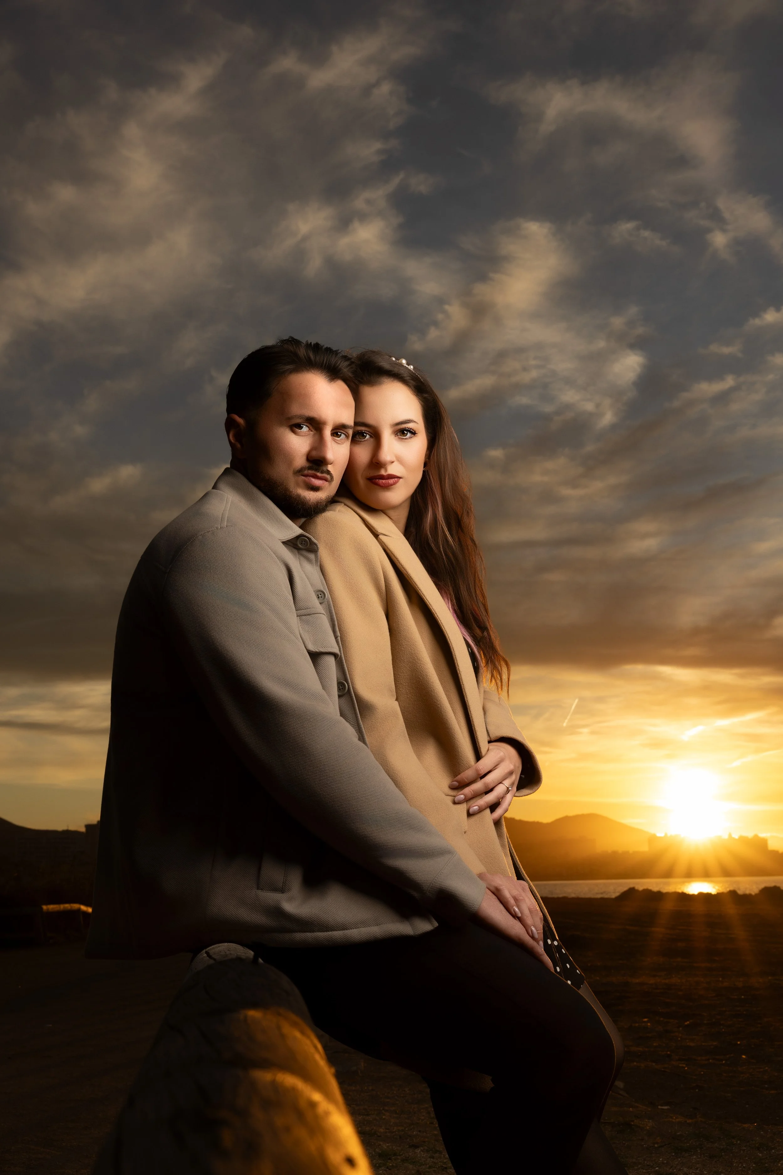 Una pareja sentada en un muelle, mirando hacia la cámara, con un atardecer en el fondo y cielo nublado.