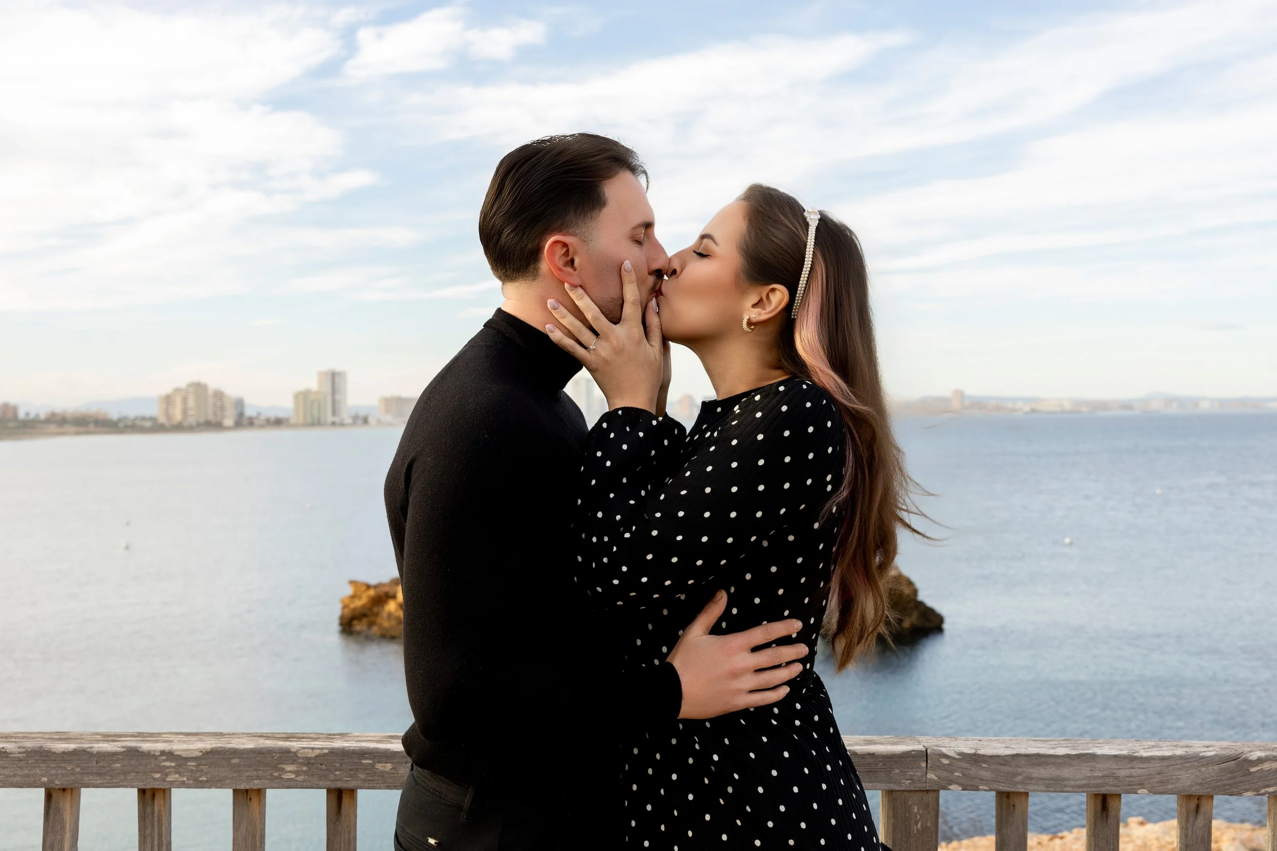 Una pareja besándose en un lugar con vista al mar y edificios en el fondo.