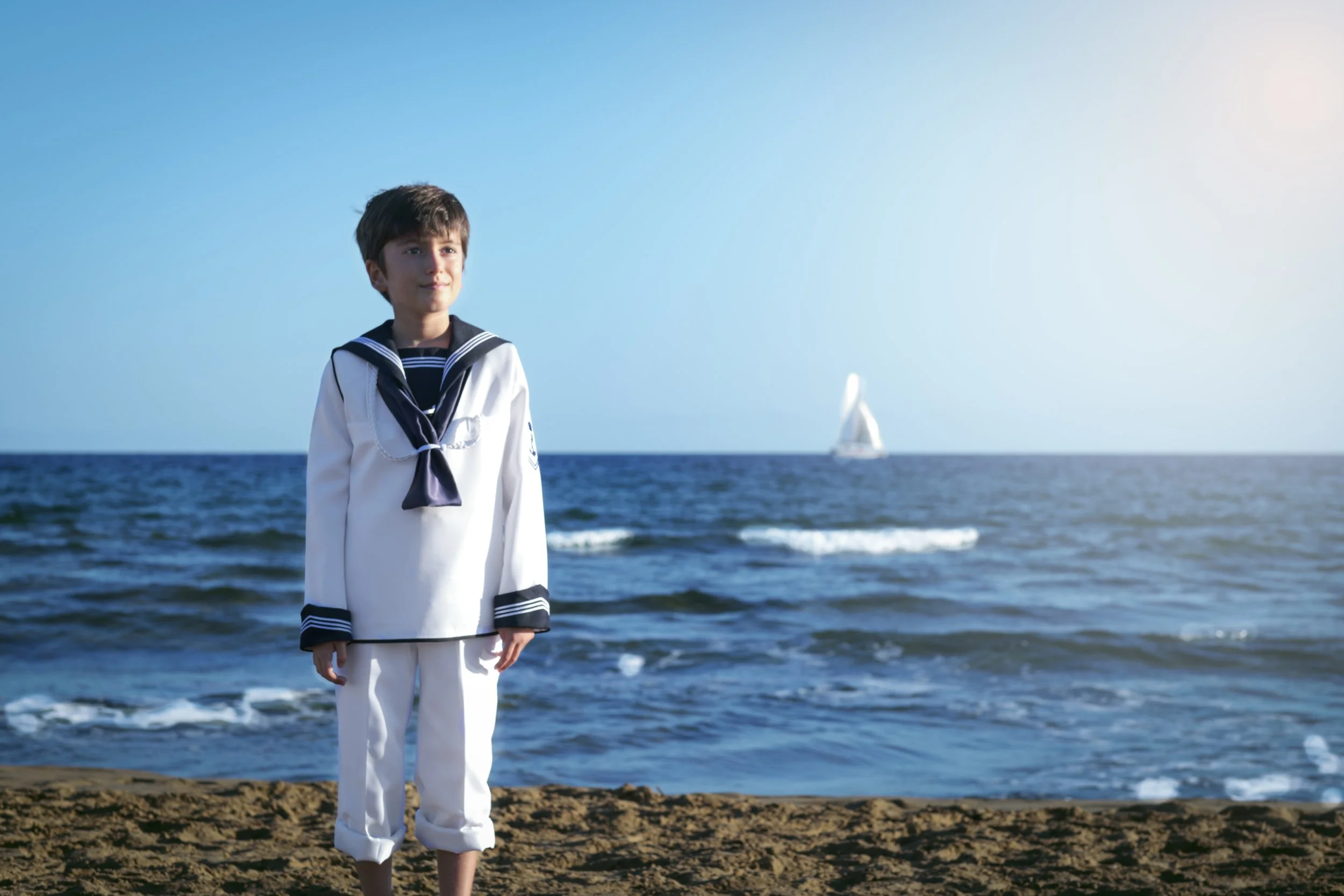 Niño vestido con uniforme escolar marinero, de pie en la arena de la playa junto al mar, con un velero en el horizonte y cielo despejado.