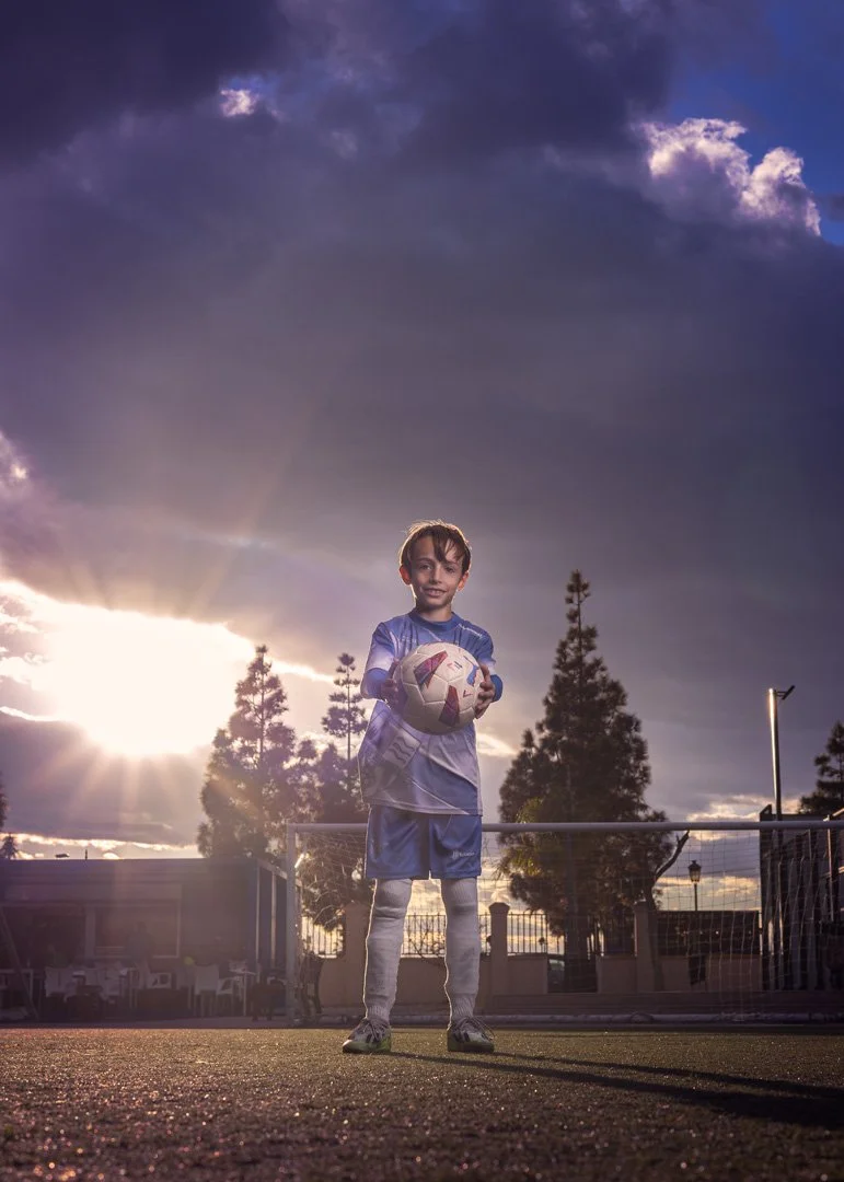 Niño con uniforme de fútbol sosteniendo un balón en un campo de fútbol, con cielo nublado y el sol poniéndose en el fondo.