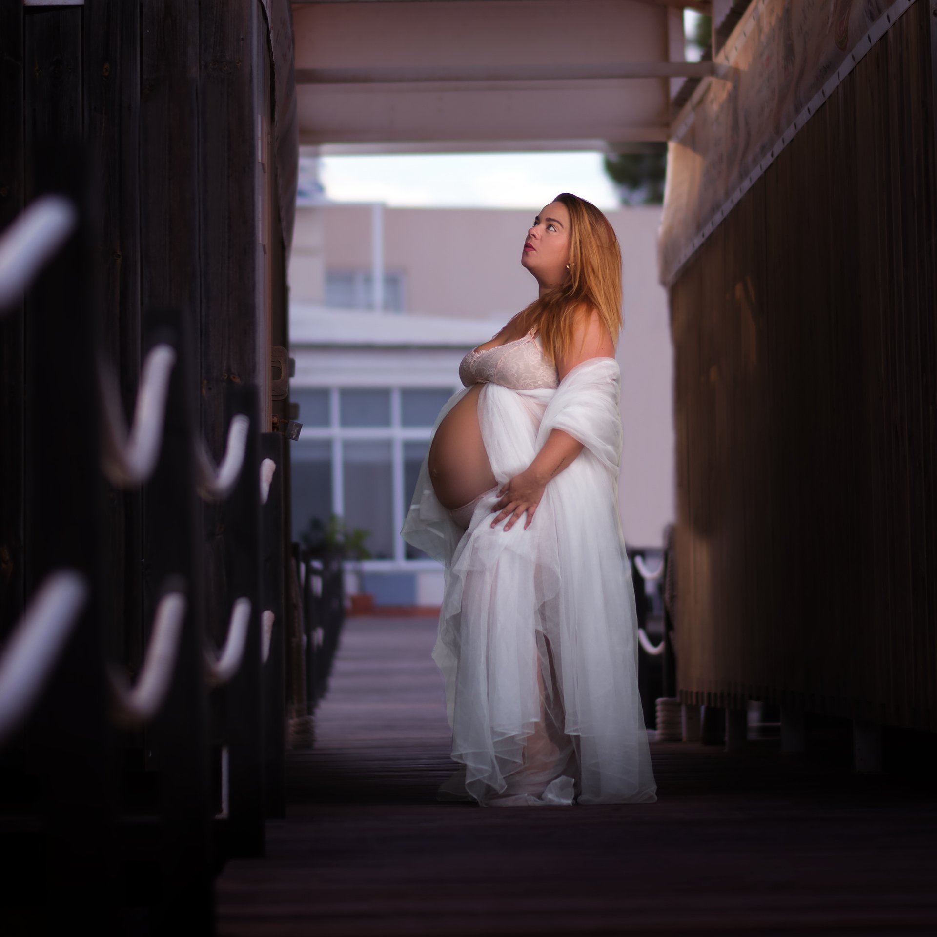 Mujer embarazada en un pasillo estilo dosel, con ropa blanca y expresión pensativa, en un entorno de exteriores con edificios en el fondo.