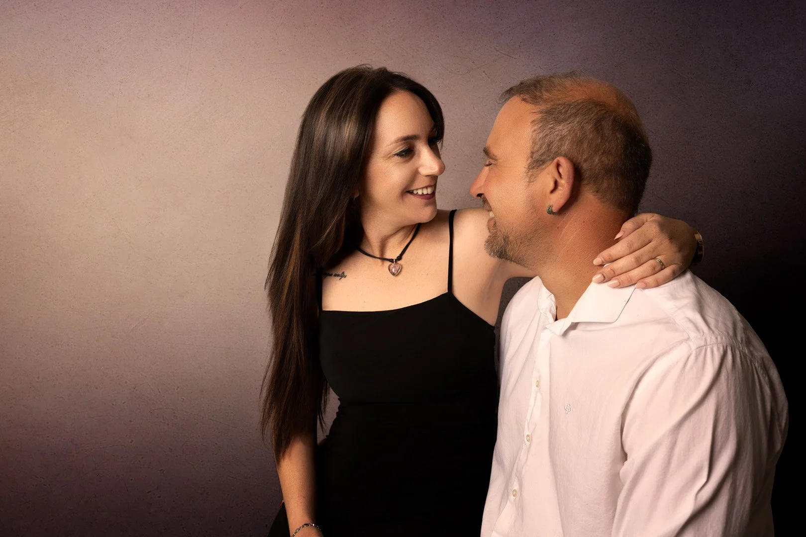 Una pareja de adultos jóvenes sonriendo y mirándose afectuosamente, ella con una mano en el hombro de él, en un fondo oscuro.