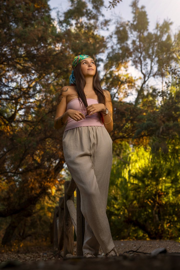 Joven mujer con bandana colorida, top rosa y pantalones beige, de cabello largo, de pie en un puente de madera en un bosque de árboles en otoño, bajo la luz del sol.
