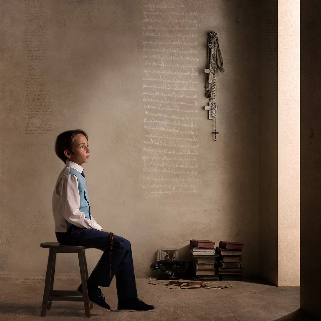 Un niño sentado en una banca en una habitación con paredes de color beige, con libros apilados en el suelo y rosarios colgados en la pared.