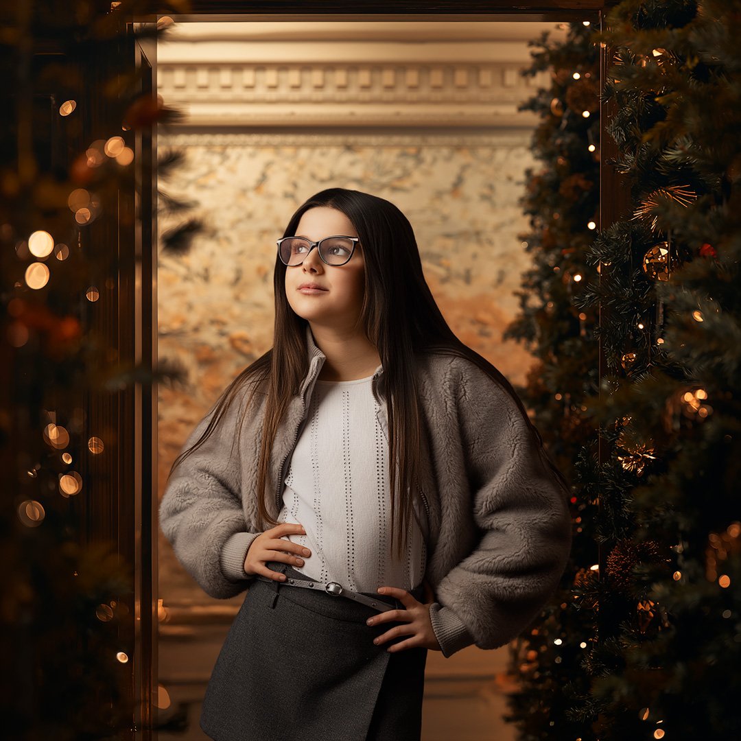Joven mujer con gafas, cabello largo y oscuro, posando entre dos árboles de Navidad decorados con luces y esferas en un ambiente cálido y festivo.