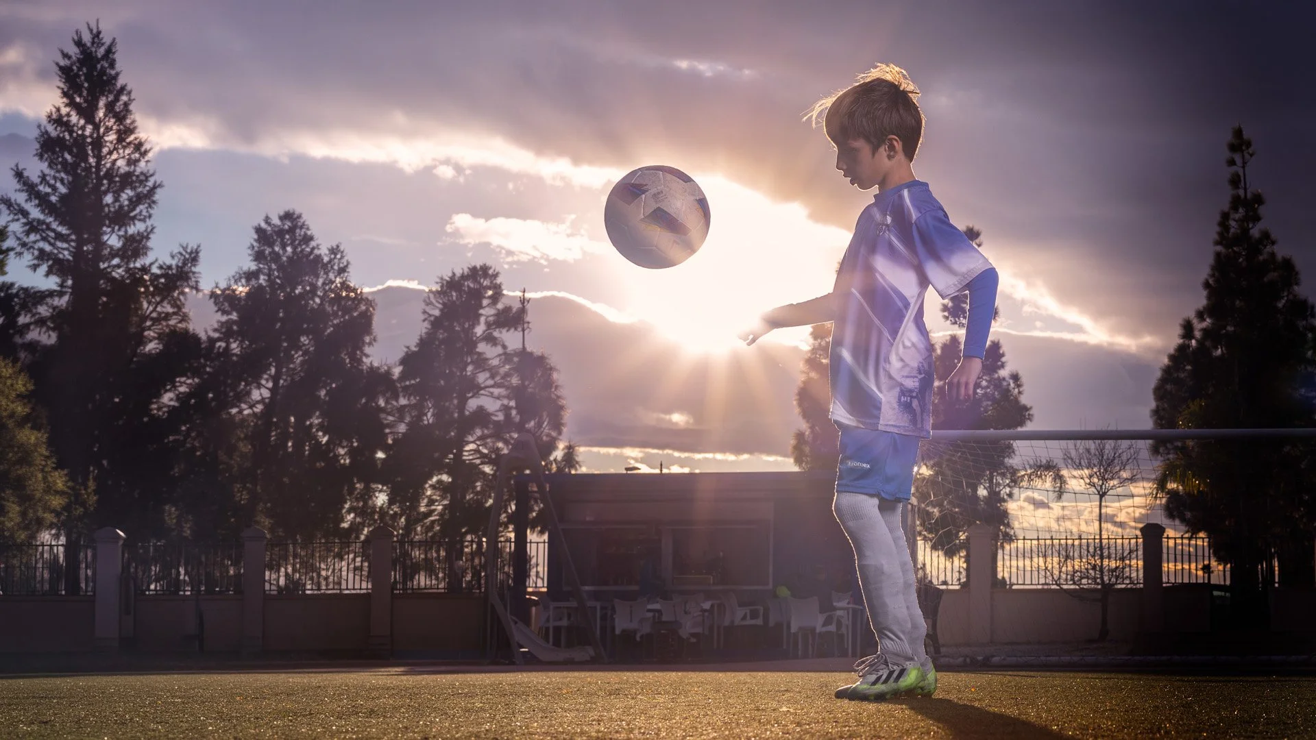Niño jugando fútbol en un campo al atardecer, con árboles y cielo nublado de fondo.