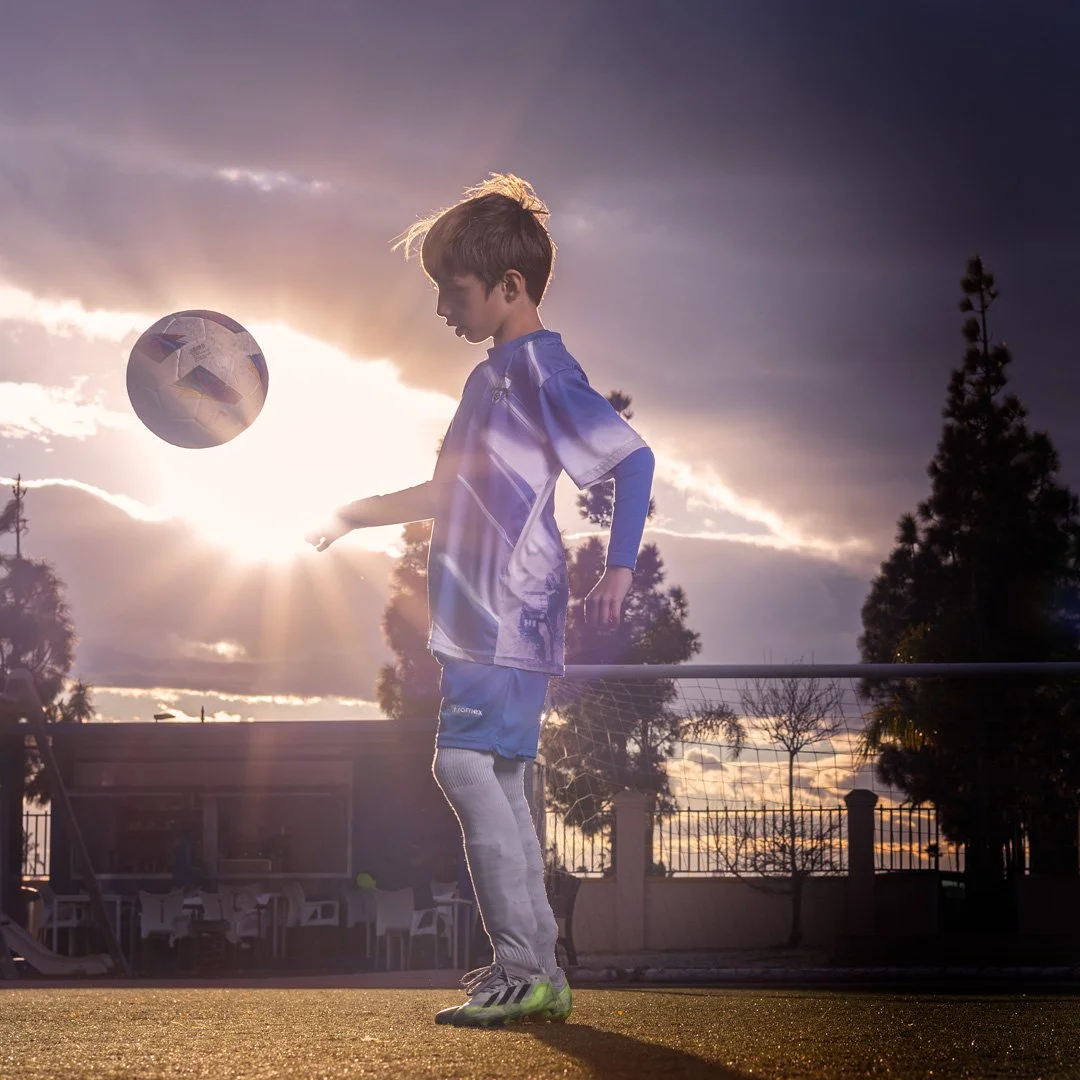 Un niño jugando fútbol en un campo al atardecer, con una pelota flotando en el aire y el sol en el fondo.