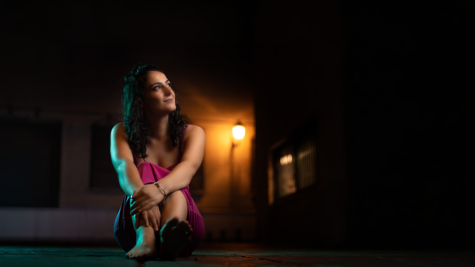 Mujer de cabello rizado sentada en el suelo, vestida con un vestido púrpura, bajo una luz cálida en un ambiente oscuro.