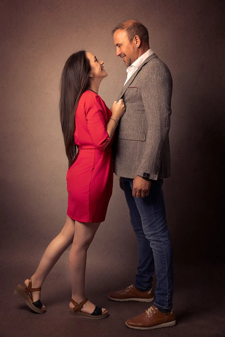 Una pareja mira con cariño, la mujer con vestido rojo y el hombre con saco gris, en un fondo neutro.