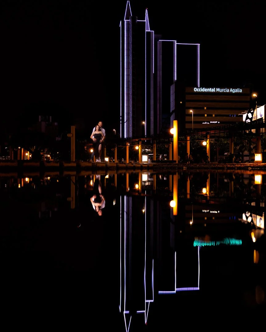 Edificio alto con iluminación púrpura y cielo oscuro, reflejado en agua, con una persona en primer plano y señalización que dice 'Occidental Murcia Agalia'.