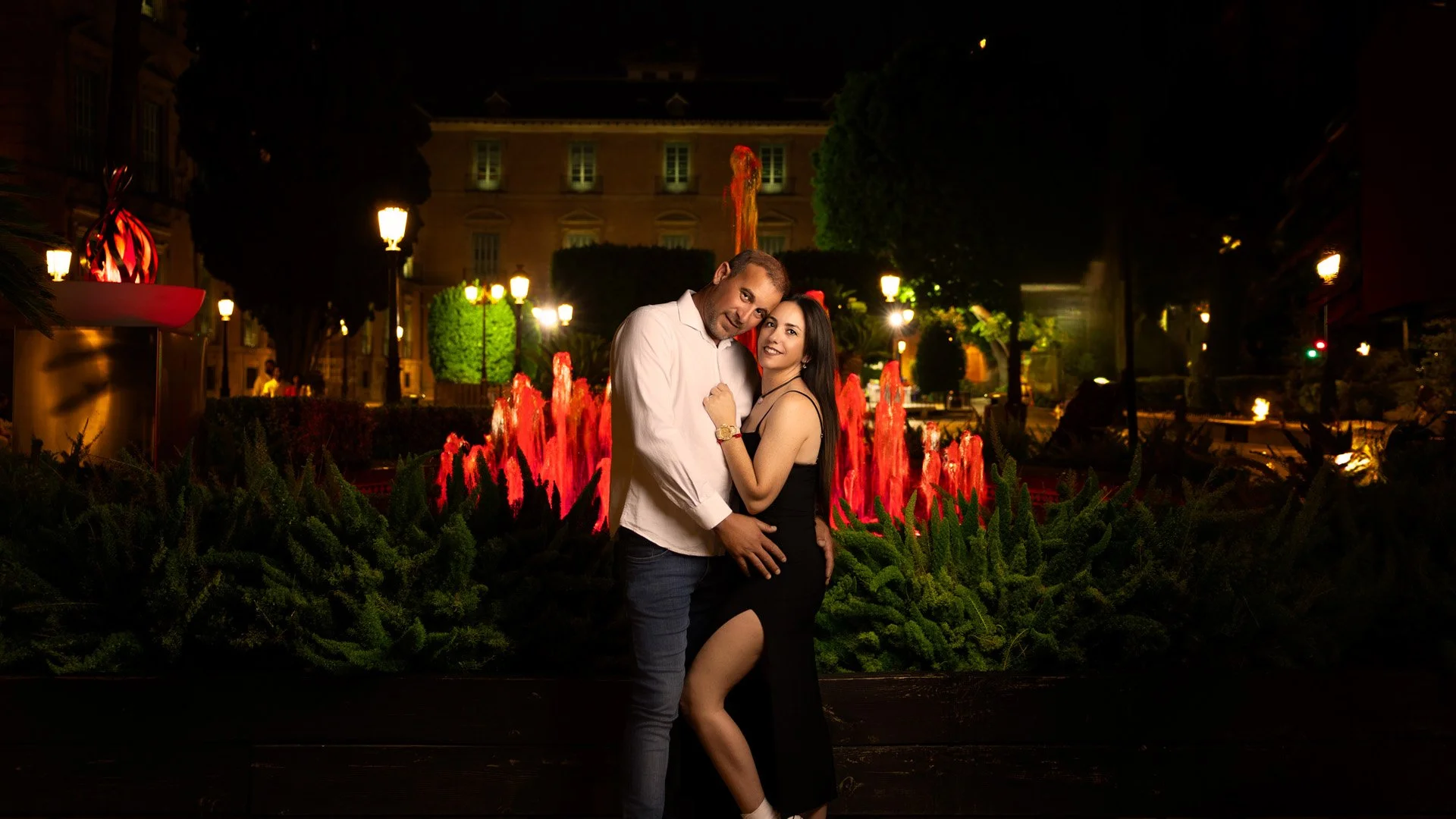 Una pareja abrazada frente a una fuente iluminada en un parque por la noche.