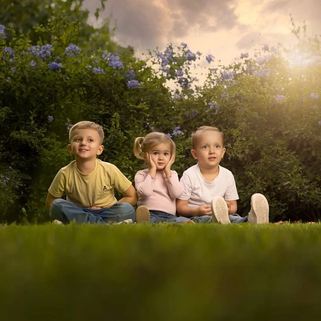Tres niños sentados en el césped, rodeados de arbustos con flores moradas, al atardecer, con un cielo con nubes y la luz del sol filtrándose.