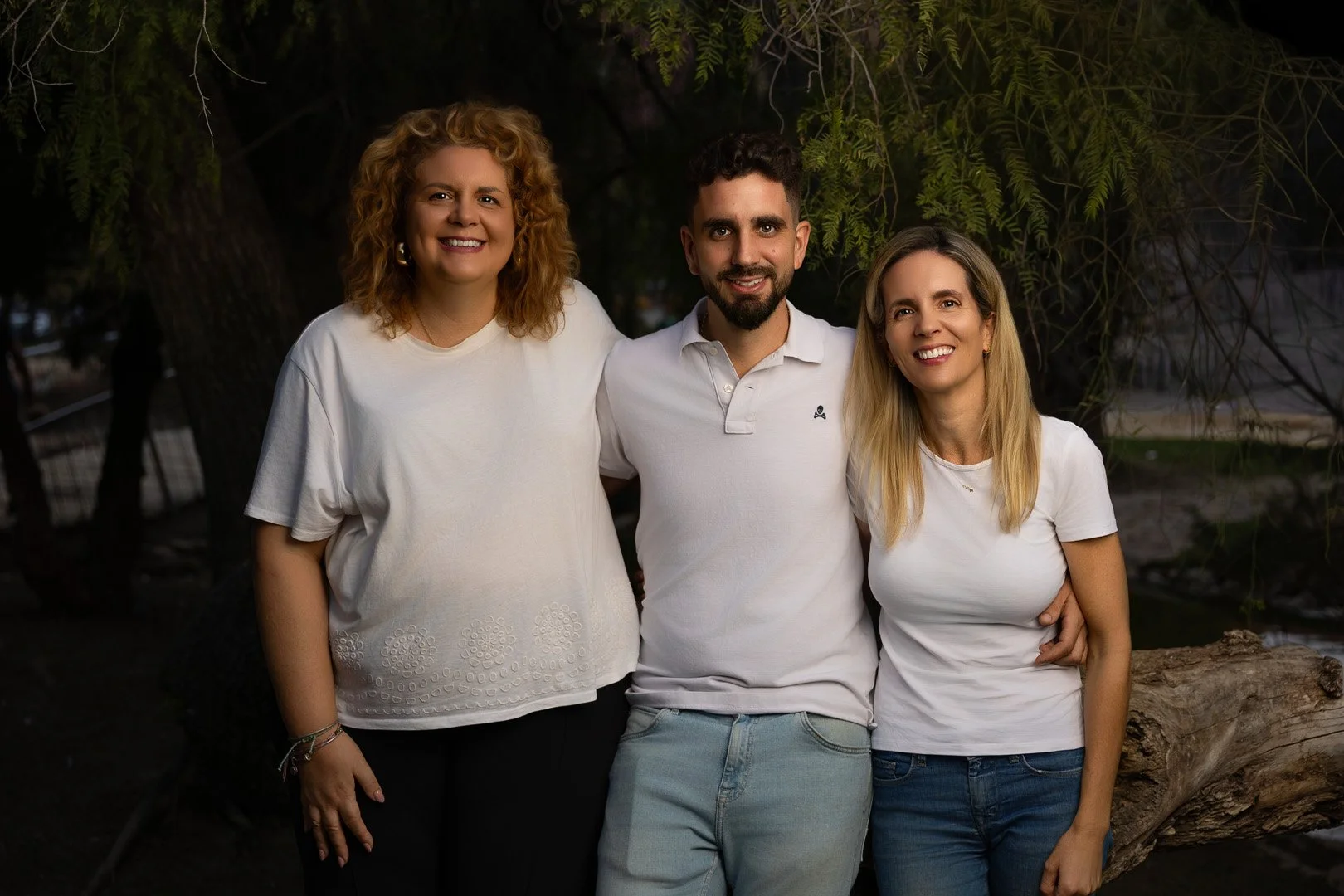 Tres personas sonrientes posando juntas al aire libre, con árboles y vegetación en el fondo.