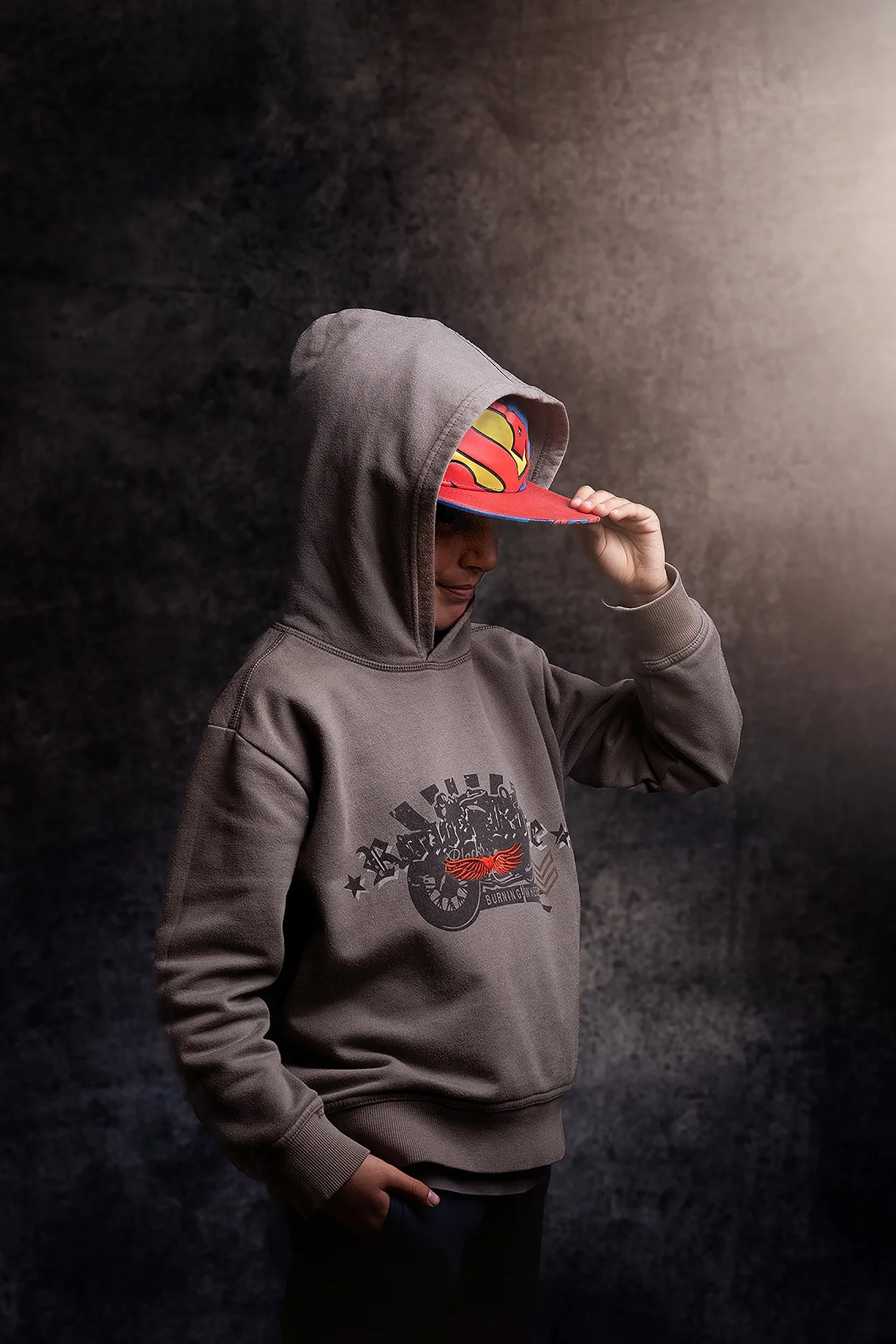 Un niño con capucha gris y una gorra de Superman se está tocando la visera con una mano, en un fondo oscuro con una luz suave a la derecha.