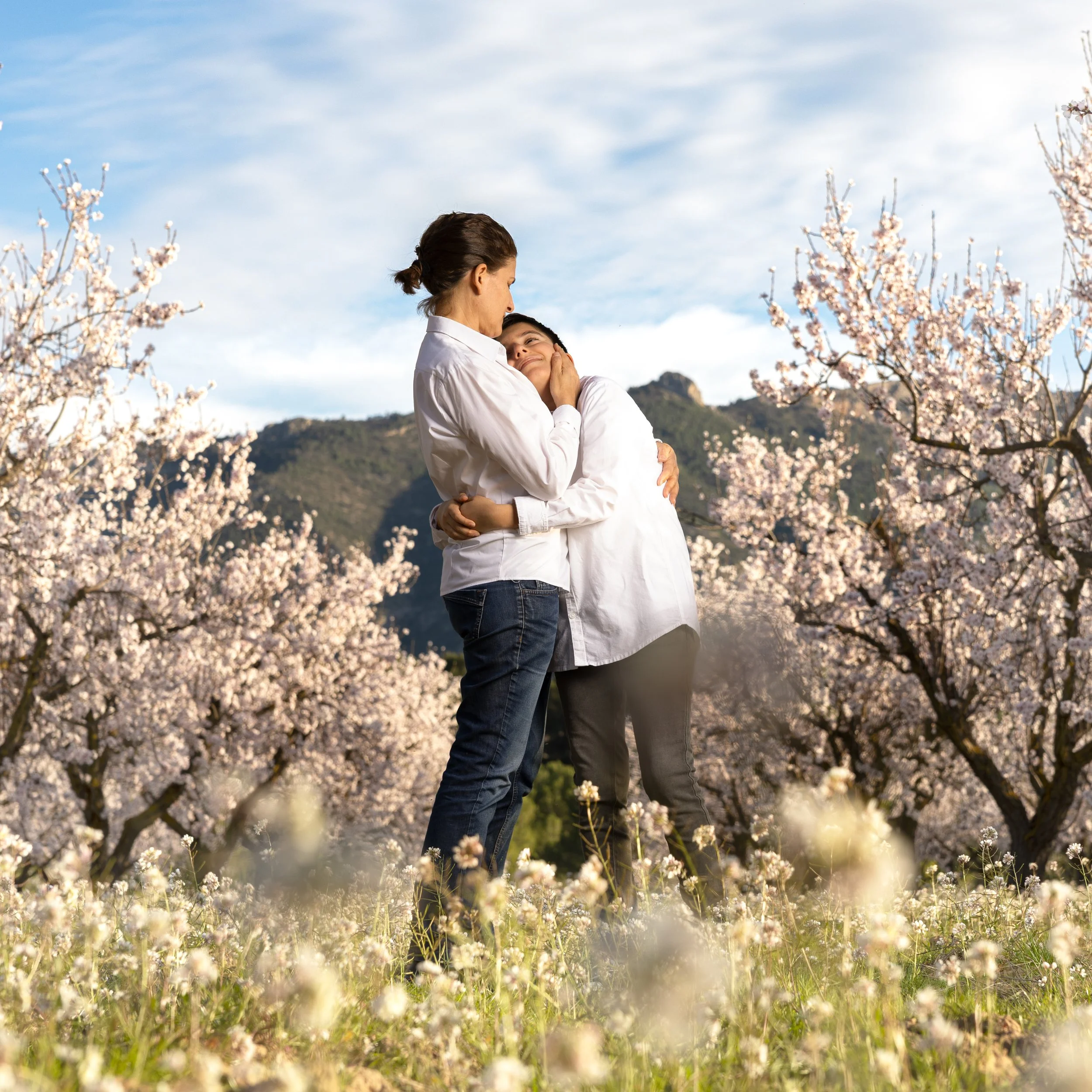 Una pareja abrazándose en un campo de flores de primavera con cerezos en flor y montañas en el fondo.