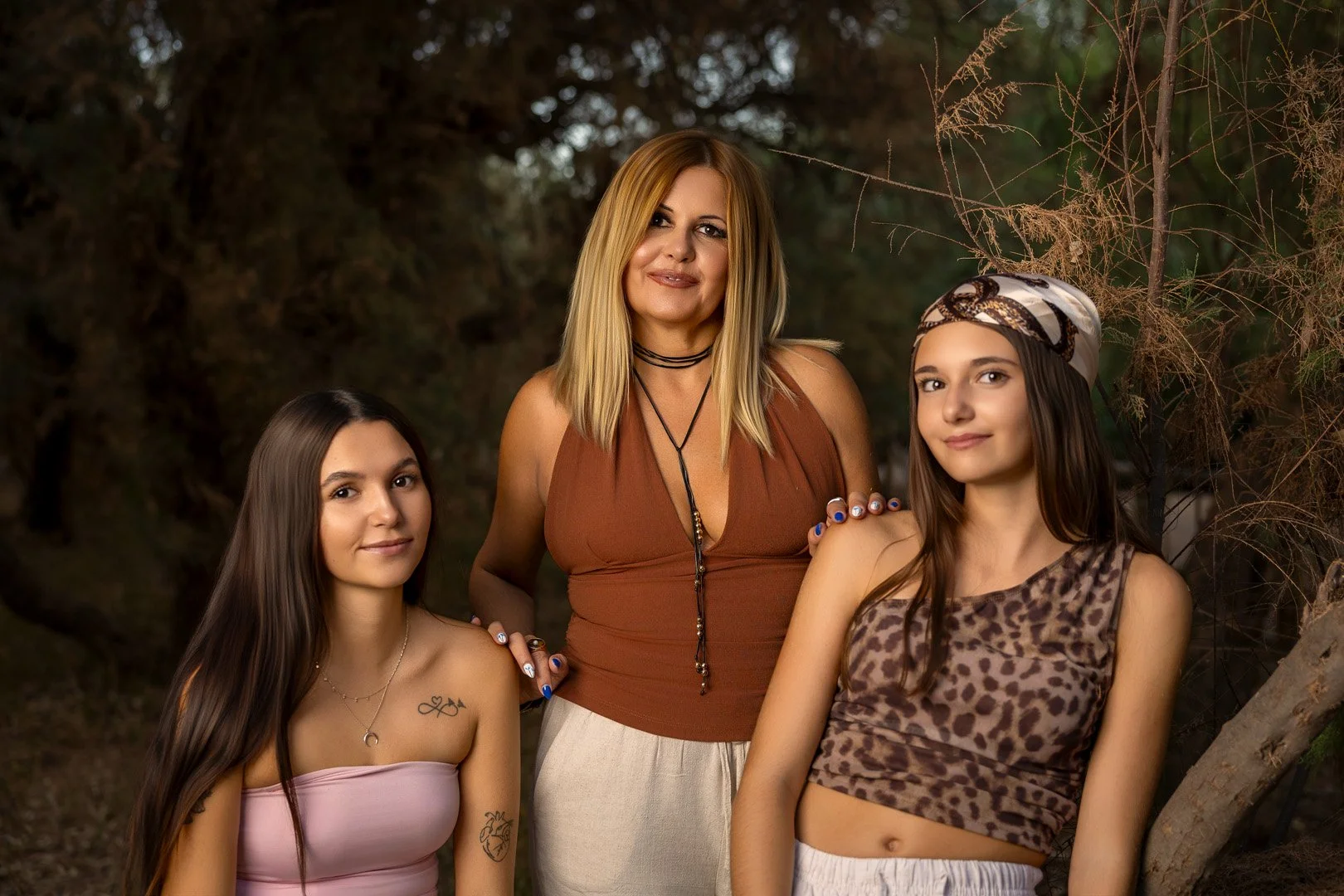 Mujer adulta con cabello rubio y dos jóvenes mujeres con cabello largo, una con tela en la cabeza. Están en un entorno natural con árboles.