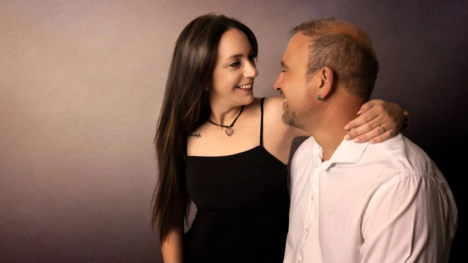 Una pareja sonriente mira con cariño y cariño, la mujer con un vestido negro y el hombre con camisa blanca, en un ambiente íntimo y afectuoso.