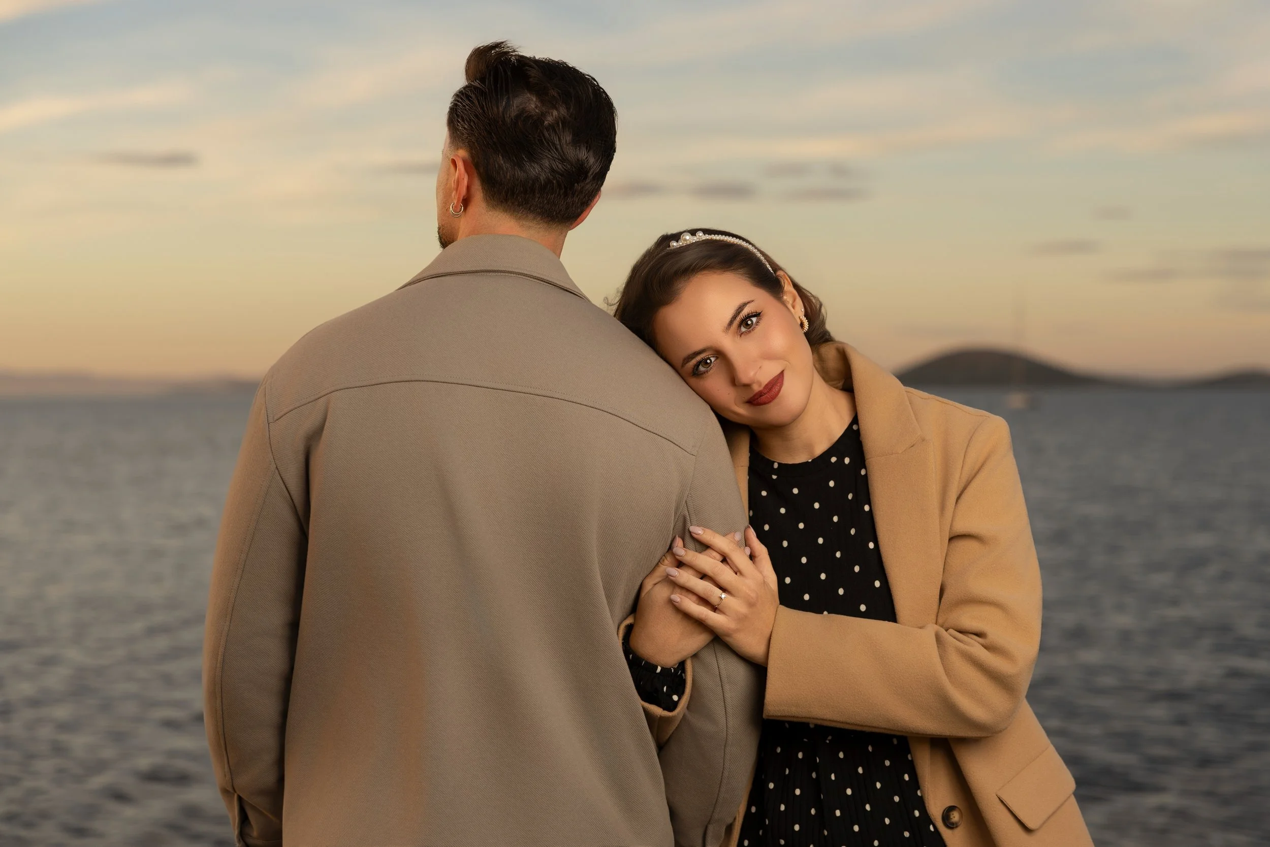 Una pareja abraza junto a un lago al atardecer, ella con vestido de lunares y él con abrigo beige.