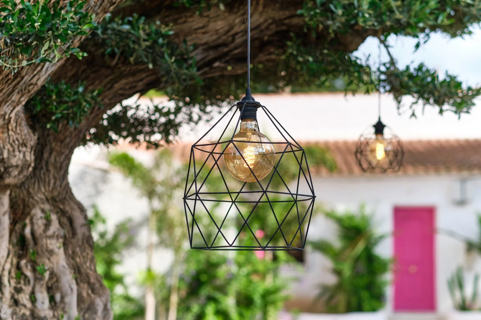 Lámpara colgante de diseño geométrico sobre un árbol en un entorno exterior con fondo de vegetación y casa con pared rosa
