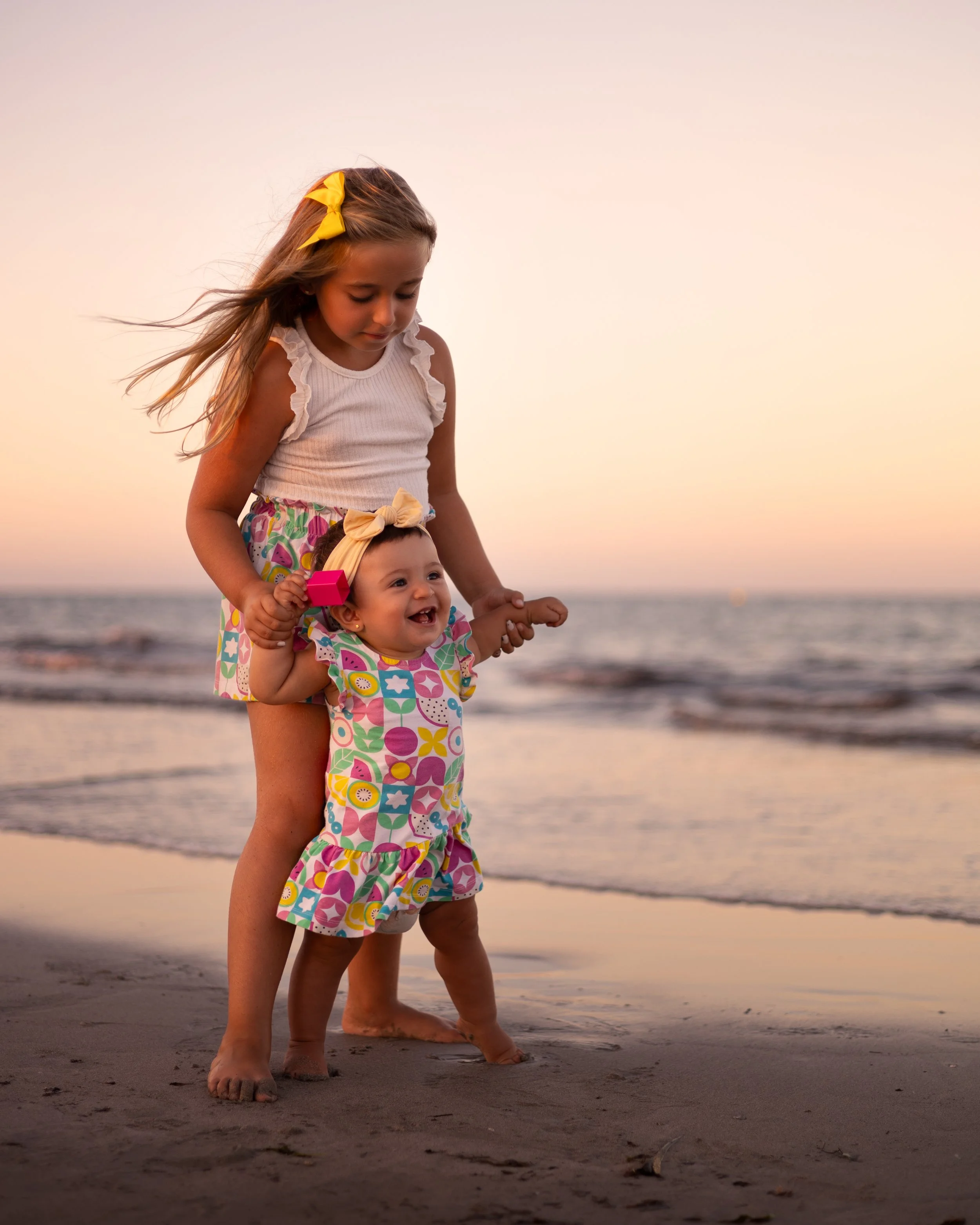 Dos niñas pequeñas jugando en la playa al atardecer, la mayor ayuda a la menor a caminar, ambas con ropa colorida y accesorios de cabeza, se ven felices y disfrutando del momento.