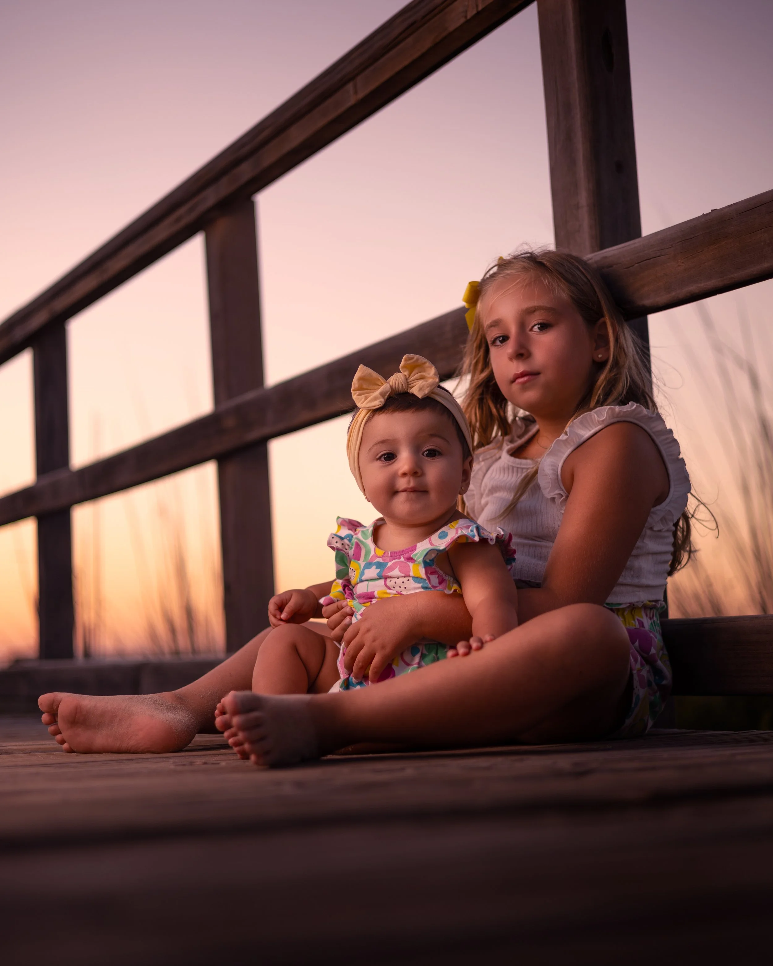 Dos niñas sentadas juntas en un muelle durante el atardecer, una niña más pequeña en brazos de la mayor, ambas con ropa colorida y expresión tranquila.