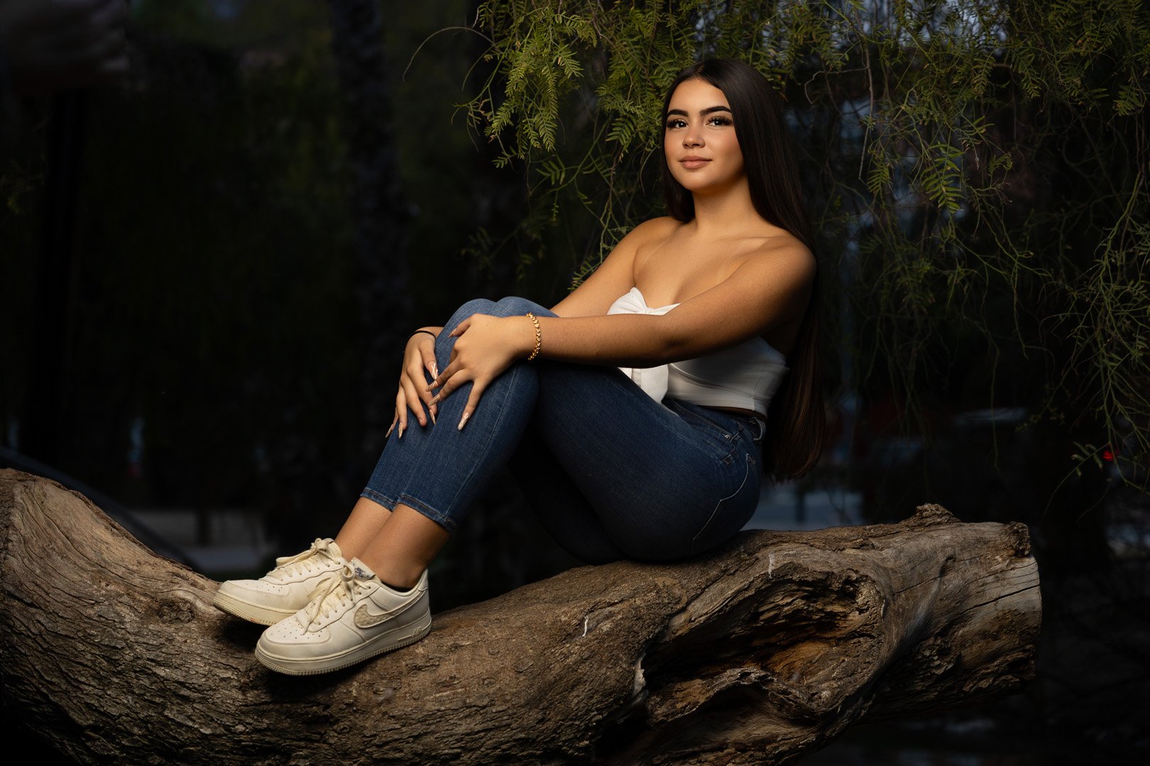 Joven mujer sentada en una rama de árbol en un entorno natural, vista de cuerpo completo, con blusa blanca, jeans ajustados, zapatillas blancas y cabello largo y oscuro.