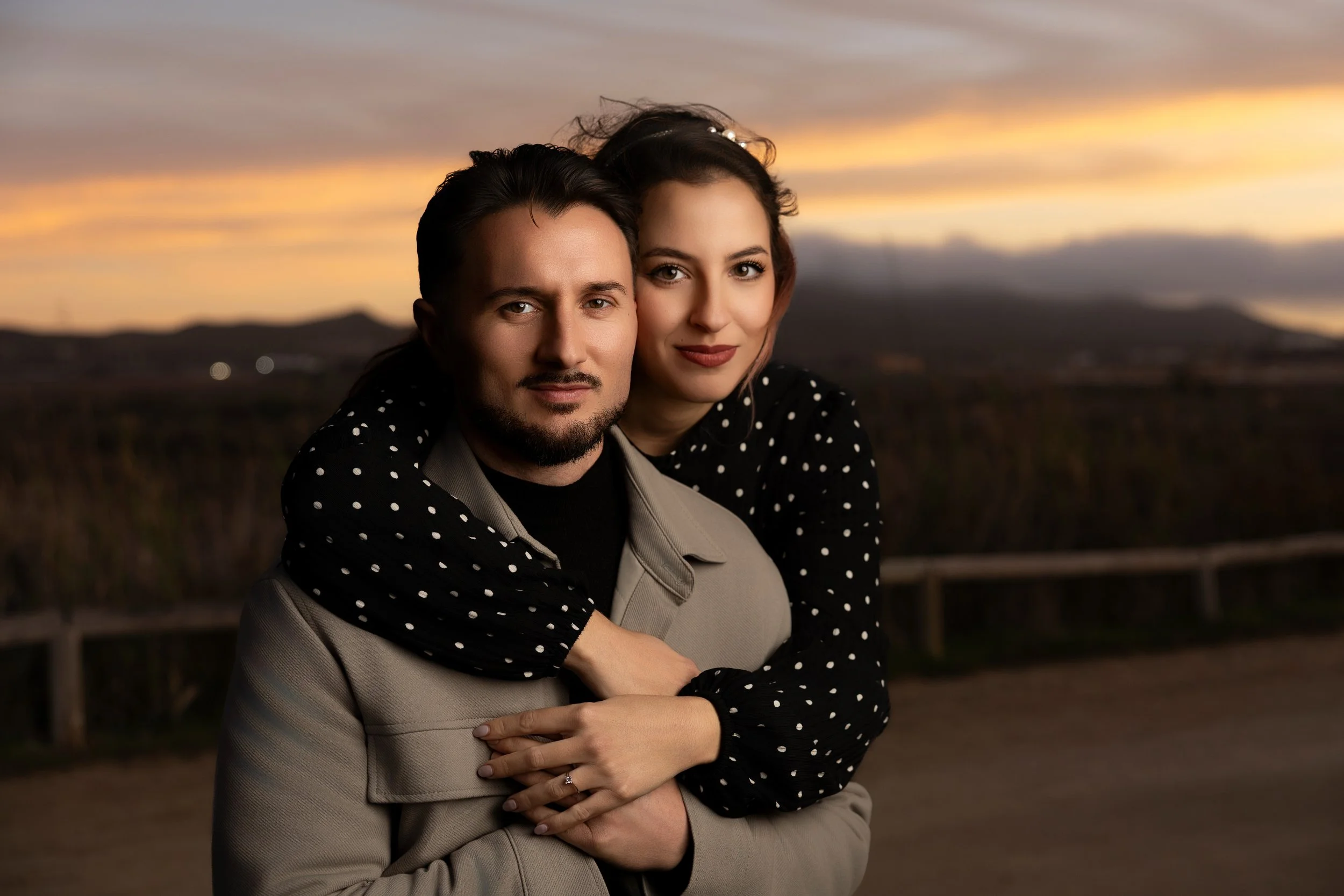 Una pareja abrazada en un paisaje al atardecer con montañas y cielo colorido