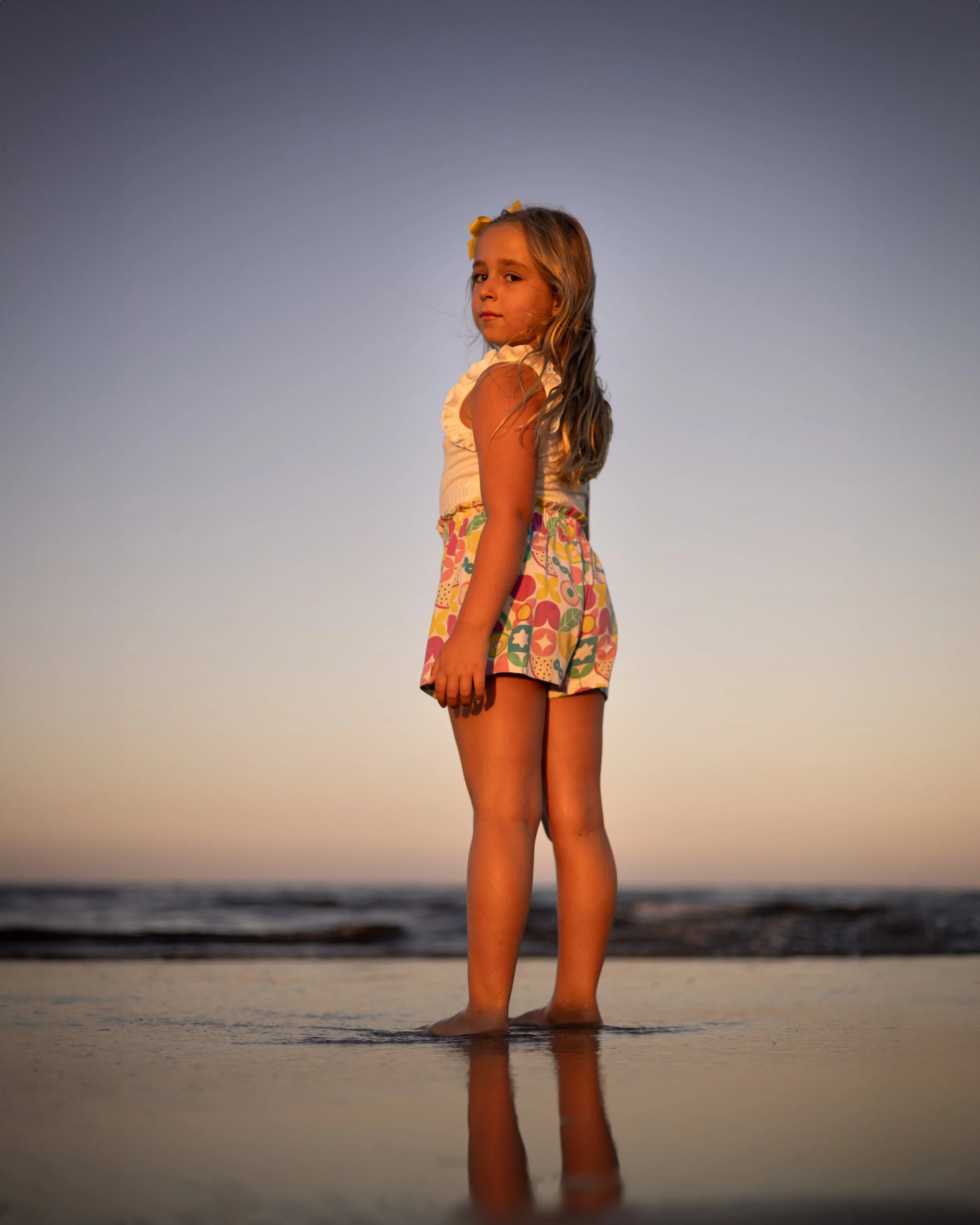 Niña de cabello rubio con lazo amarillo en un día soleado en la playa, de pie en la arena con agua hasta los tobillos, mirando hacia la cámara con fondo del mar y cielo despejado.