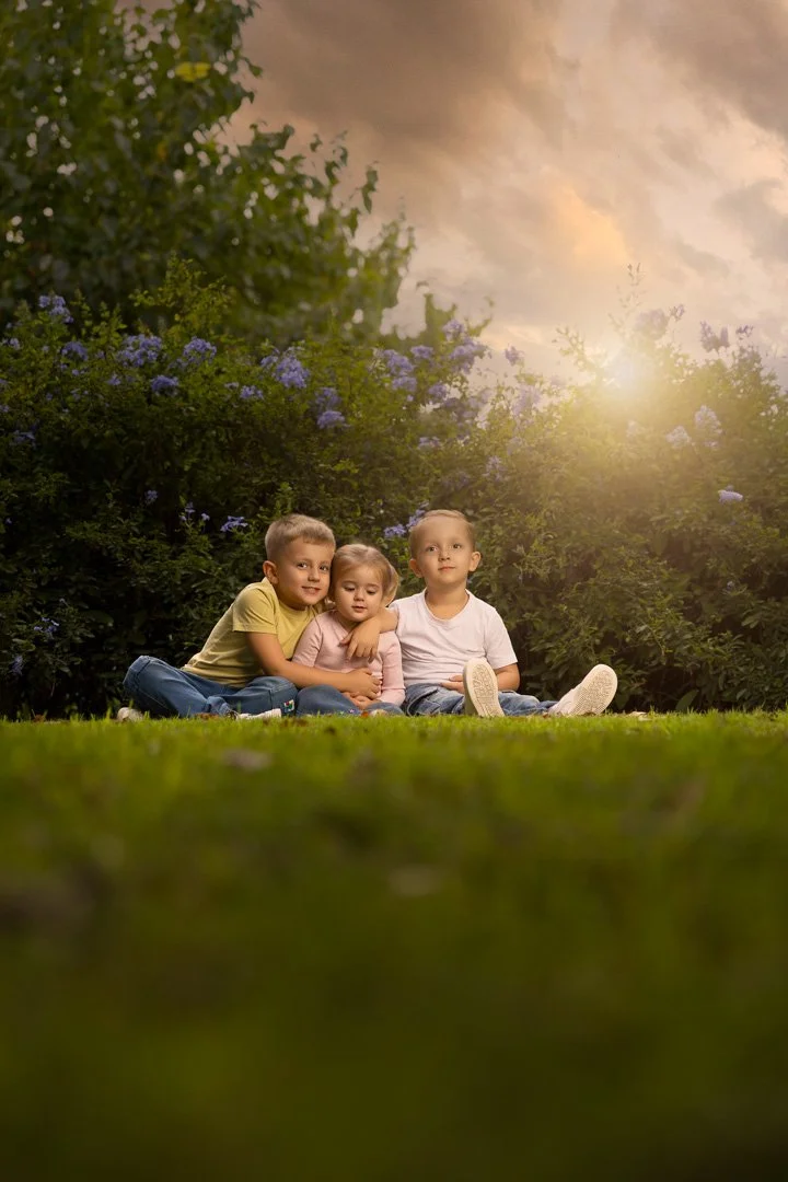 Tres niños sentados en el césped, rodeados de arbustos con flores moradas, al atardecer.