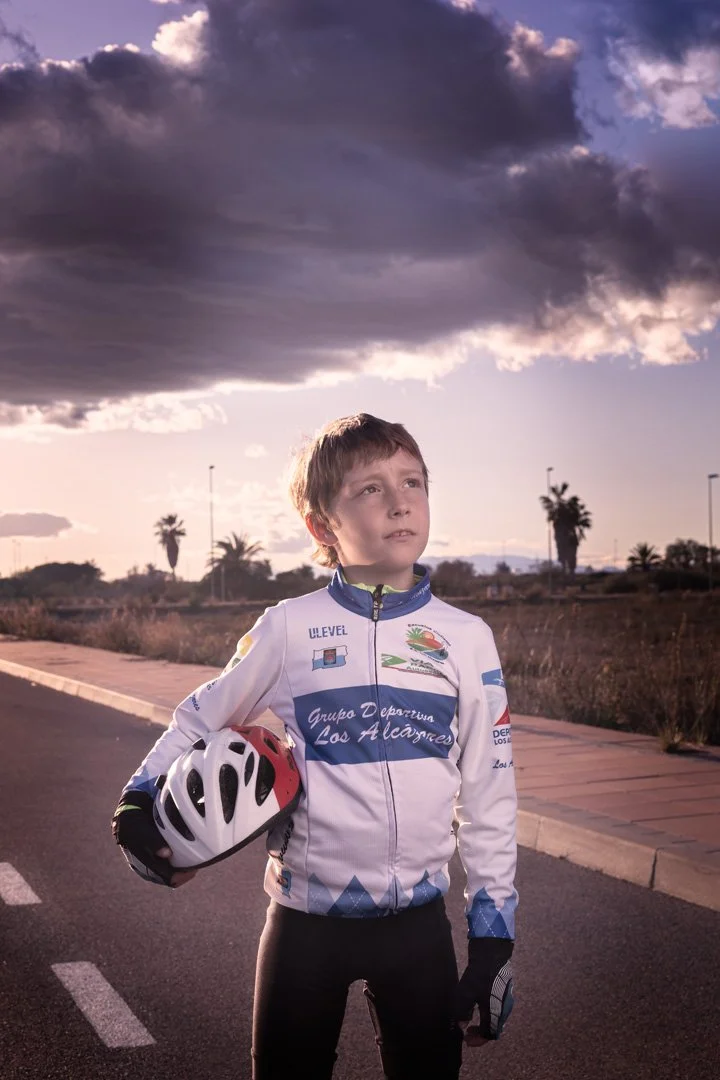 Joven ciclista con casco en la mano, vistiendo ropa deportiva, mirando hacia el cielo nublado en un entorno al aire libre con palmeras y un camino pavimentado en el fondo, al atardecer.