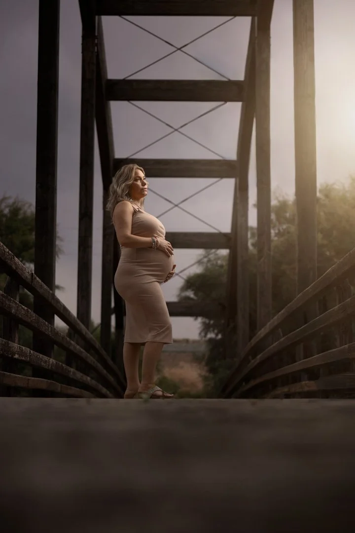 Mujer embarazada de pie en un puente de madera, en un entorno natural al atardecer.