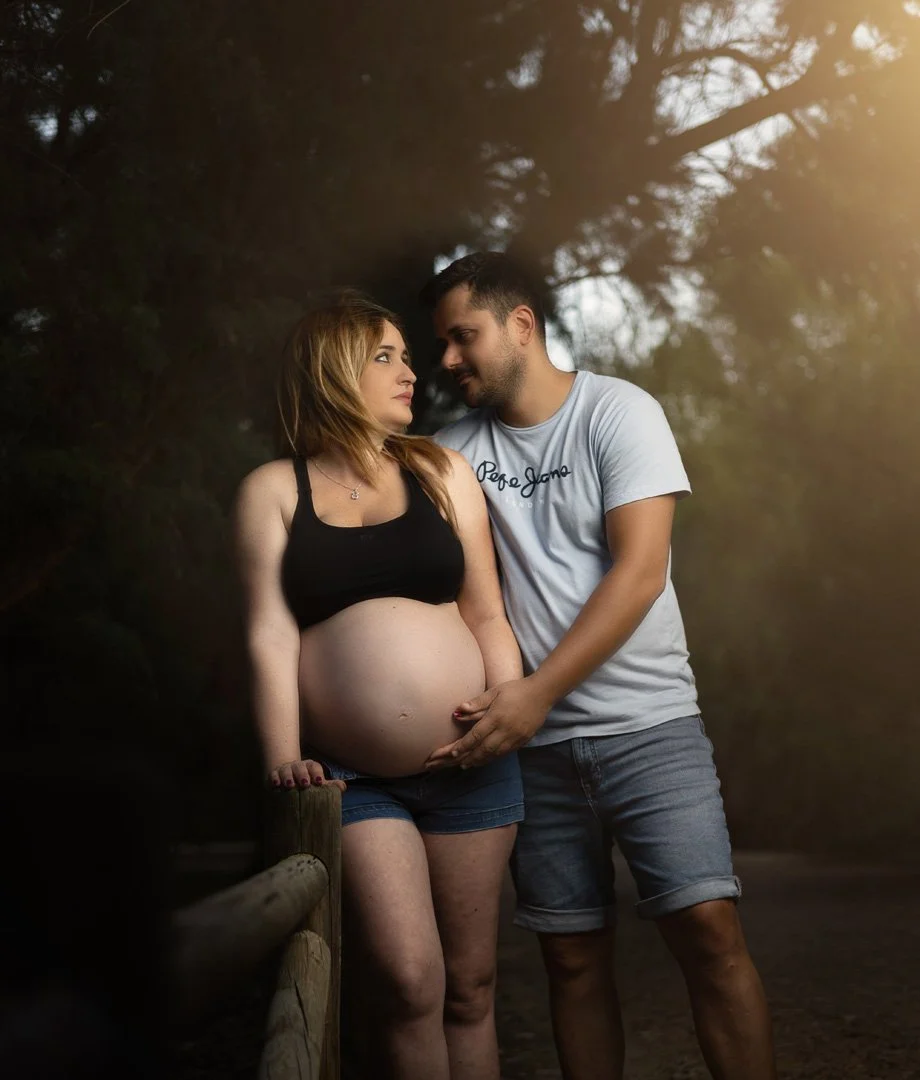 Una pareja de pie en un entorno natural, la mujer embarazada con pantalones cortos negros y una blusa negra, el hombre con camiseta gris y pantalones cortos cortos, ambos se miran con ternura en un día soleado.